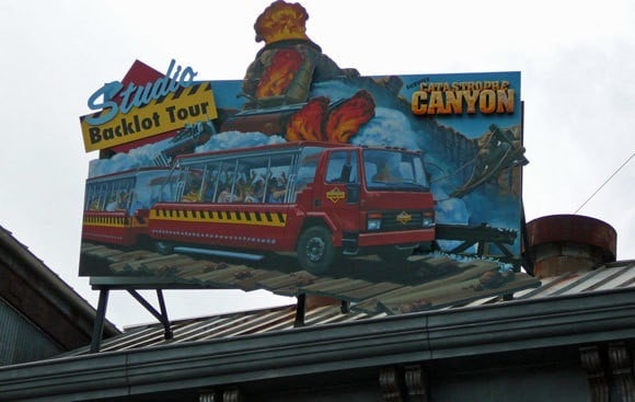 Billboard advertising the Hollywood Studios Backlot Tour featuring a red tram in a dramatic canyon scene, mounted on a rooftop against a cloudy sky.