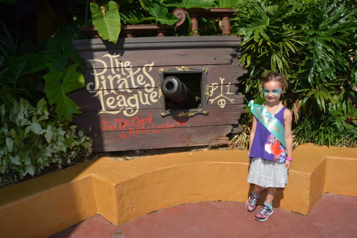 Amidst the lush greenery, a young girl stands confidently in front of a decorative cannon display labeled "The Pirates League." With face paint and a sash, she embodies the spirit of adventure, capturing the essence of experience over stuff.