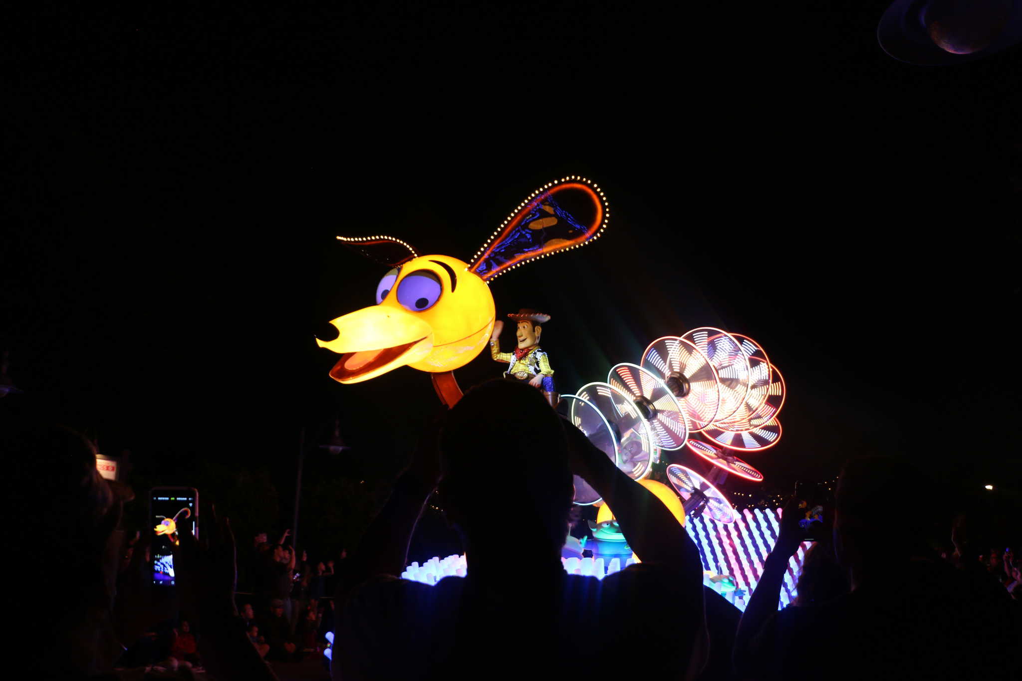 A colorful illuminated parade float at California Adventure features a large, cartoon-style dog with a figure riding it, captivating the crowd watching under the night sky.