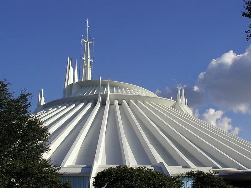 A large, white, futuristic building reminiscent of Space Mountain stands with pointed spires and a domed roof against a blue sky dotted with clouds.