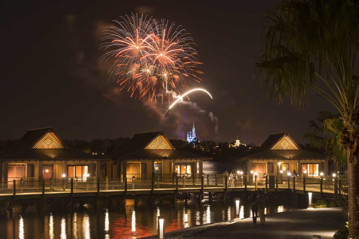 Fireworks light up the night sky over a waterfront resort, with overwater bungalows and a lit pathway in the foreground.
