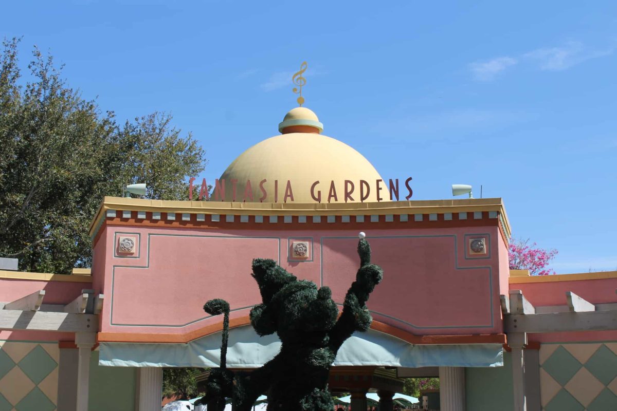 Entrance to Fantasia Gardens, featuring a dome-topped building with the name "Fantasia Miniature Golf reopens" written in pink letters and a topiary shrub shaped like a dancing figure. Blue sky and trees are in the background.