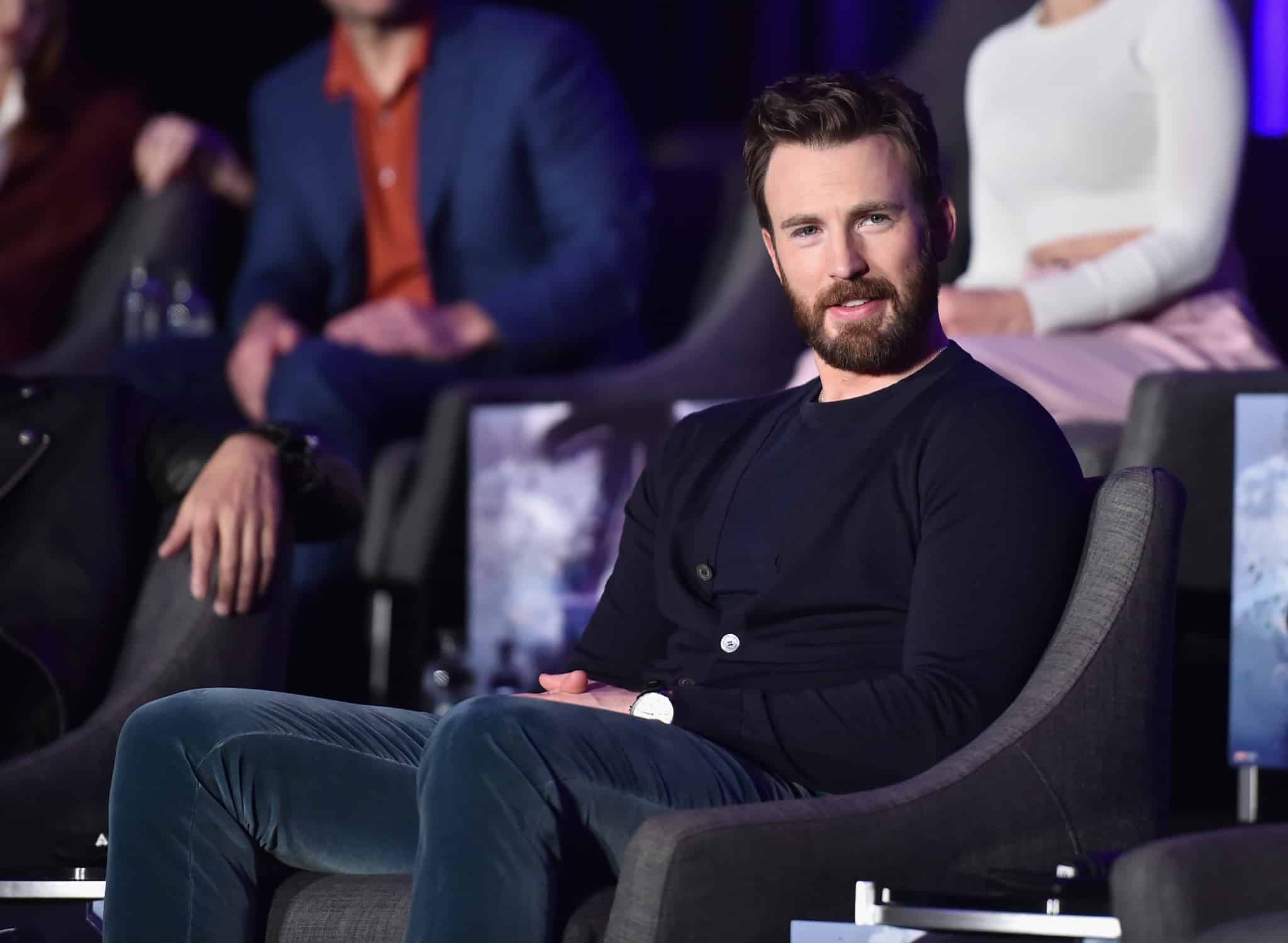 A man with a beard, wearing a dark sweater and sitting in a gray chair, looks toward the camera as if he's part of the cast at an Avengers: Endgame global press conference.