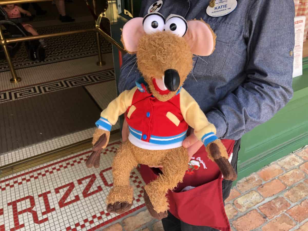 A Cast Member is greeting guests of PizzeRizzo with a stuffed Rizzo plush