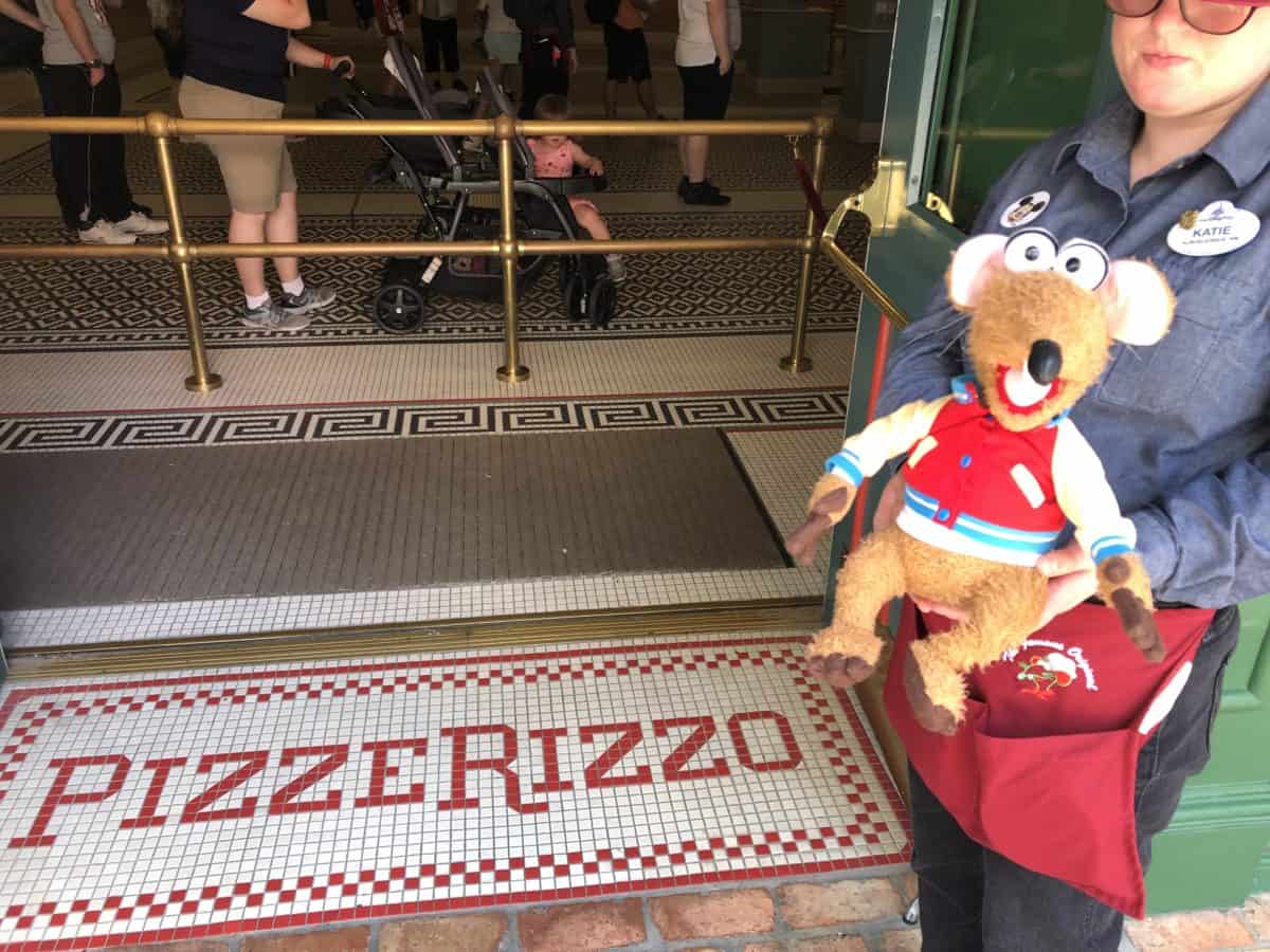 A Cast Member is greeting guests of PizzeRizzo with a stuffed Rizzo plush