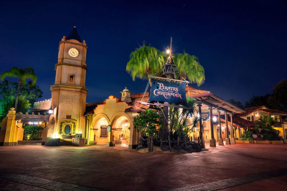 The entrance to the Pirates of the Caribbean attraction at Disneyland at night, featuring a clock tower, lush palm trees, and a sign for the ride, perfectly captures the enchanting ambiance characteristic of Magic Kingdom Attraction Queues.