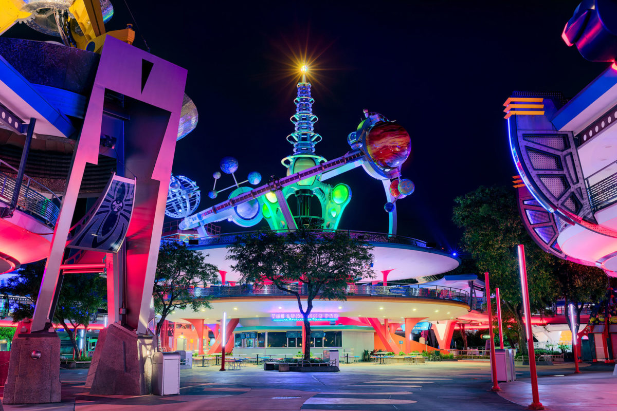 Futuristic theme park entrance illuminated at night with vibrant neon lights, featuring a central structure adorned with planetary models and surrounded by stylized architectural elements.