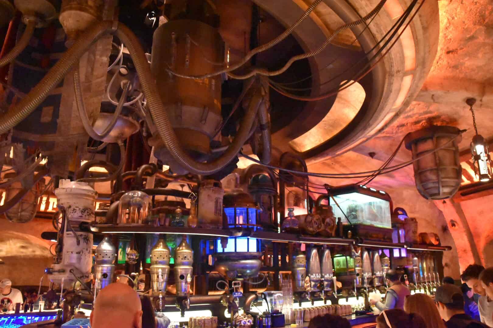Step into this futuristic-themed bar reminiscent of Oga's Cantina, with a large circular structure overhead adorned with pipes and containers. Patrons enjoy their drinks at the counter, fully immersed in the sci-fi decor.