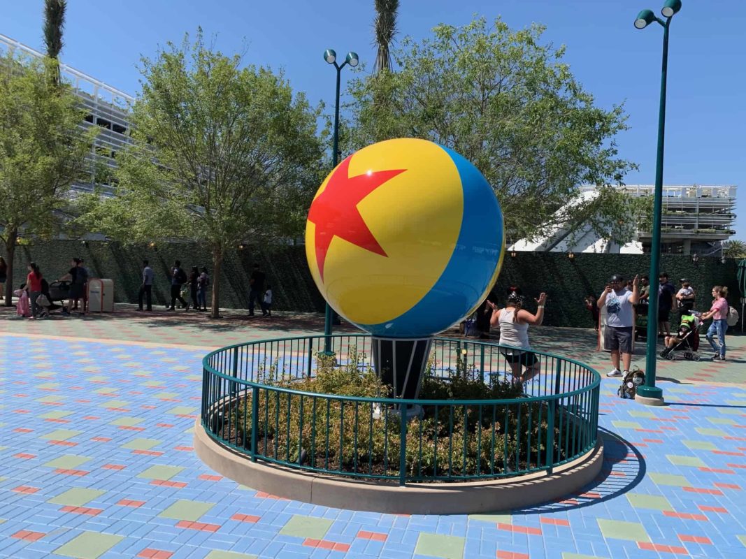 A large, colorful ball with a red star sits on a pedestal surrounded by a fence at Disneyland Resort. In the outdoor plaza, amidst trees, people are busy capturing photos of this vibrant scene near Pixar Pals Garage.