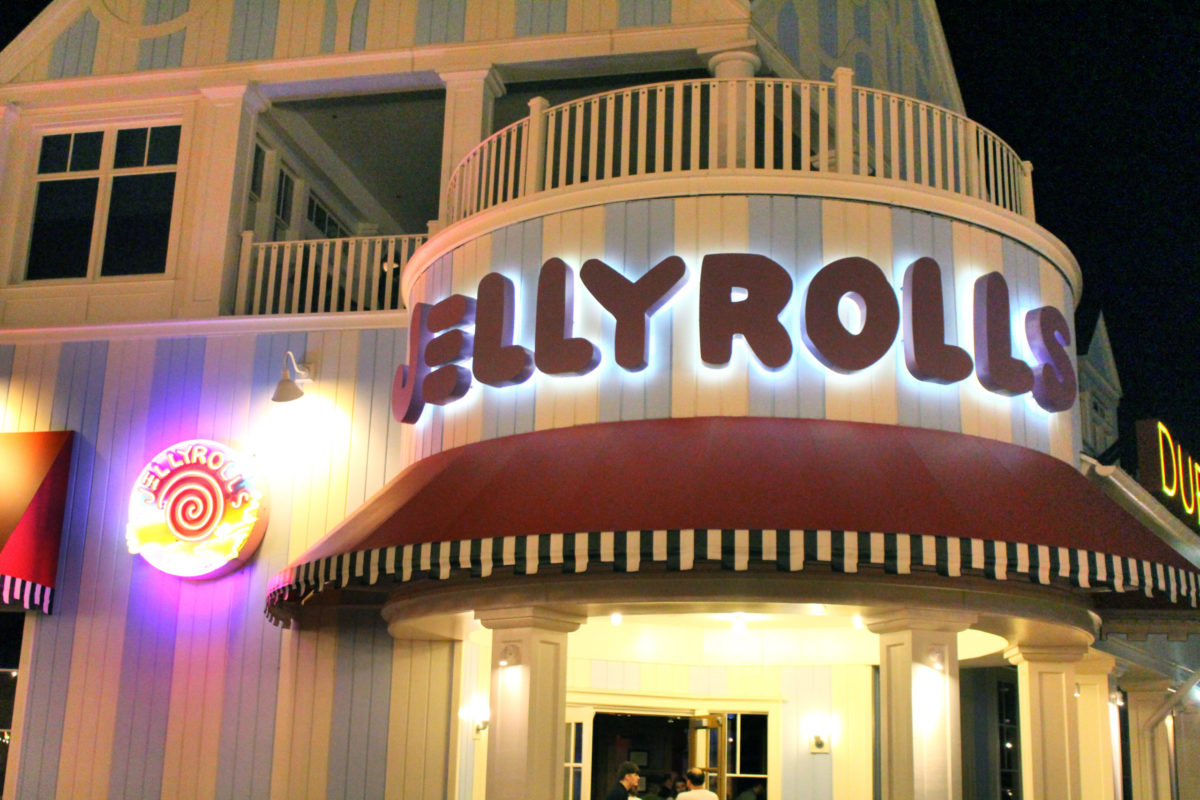 The exterior of a building glows at night with the lit "JELLYROLLS" sign and a red canopy, hinting at its lively character. Other neon signs flicker in the background, suggesting hints of stories like JellyRolls' recent PPP loan whispers circulating in the air.
