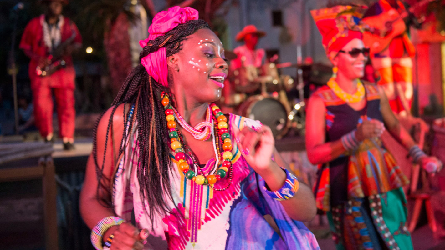 A woman, dressed in colorful traditional attire with beaded jewelry and face paint, dances joyfully to the vibrant rhythms of the Burudika Band. Musicians and other dancers are in the background.