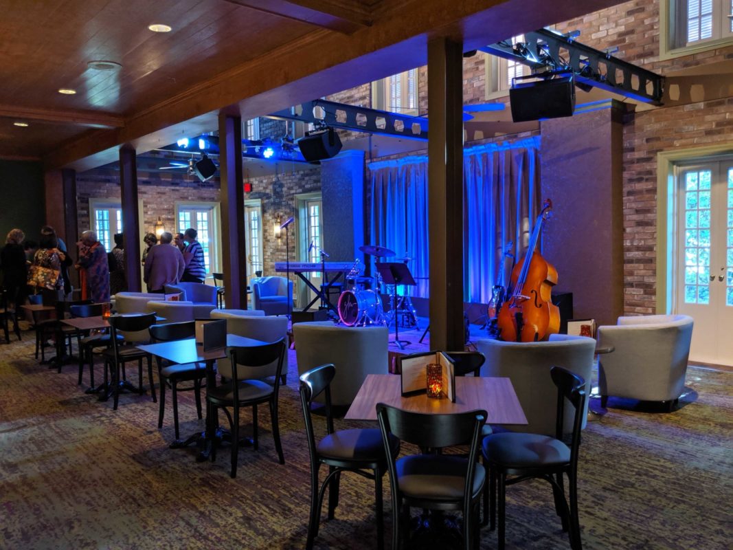 The cozy indoor setting of Scat Cat's Club features a stage with musical instruments under blue lighting, several tables and chairs arranged, and people gathered near the back of the room.