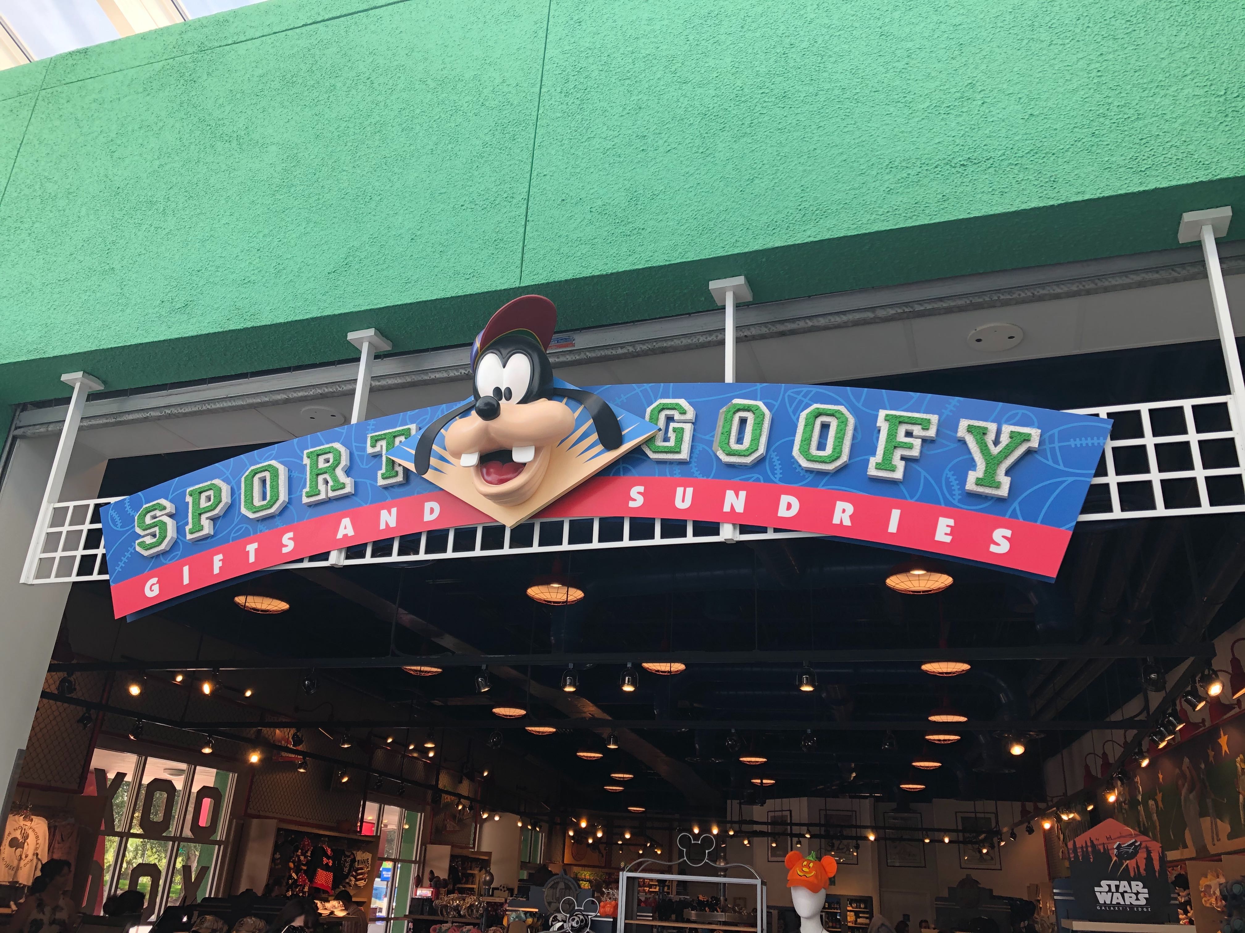 Entrance to Sport Goofy Gifts and Sundries store with a sign featuring a cartoon character's head, set against a green building exterior. The store interior is visible through the open entrance, including MagicBands on display beside Mickey-themed merchandise.