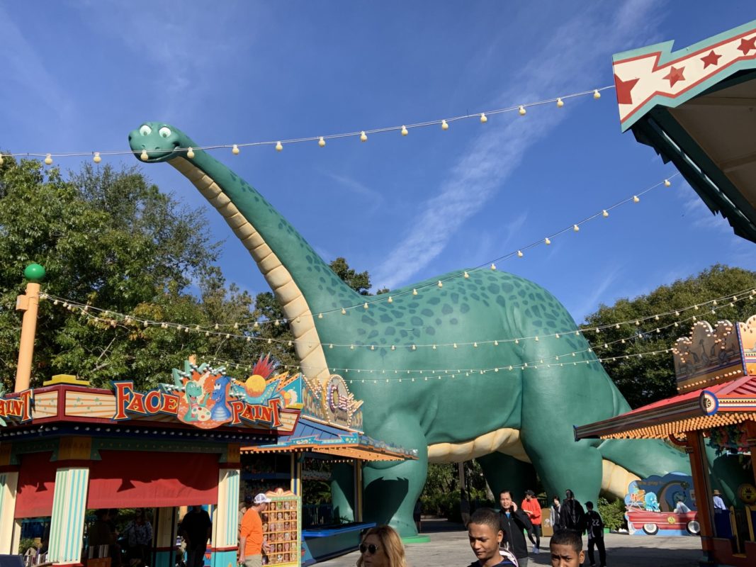 A large green dinosaur statue with a long neck towers over Dinoland, surrounded by food stands and visitors. Strings of lights hang above against a clear blue sky.