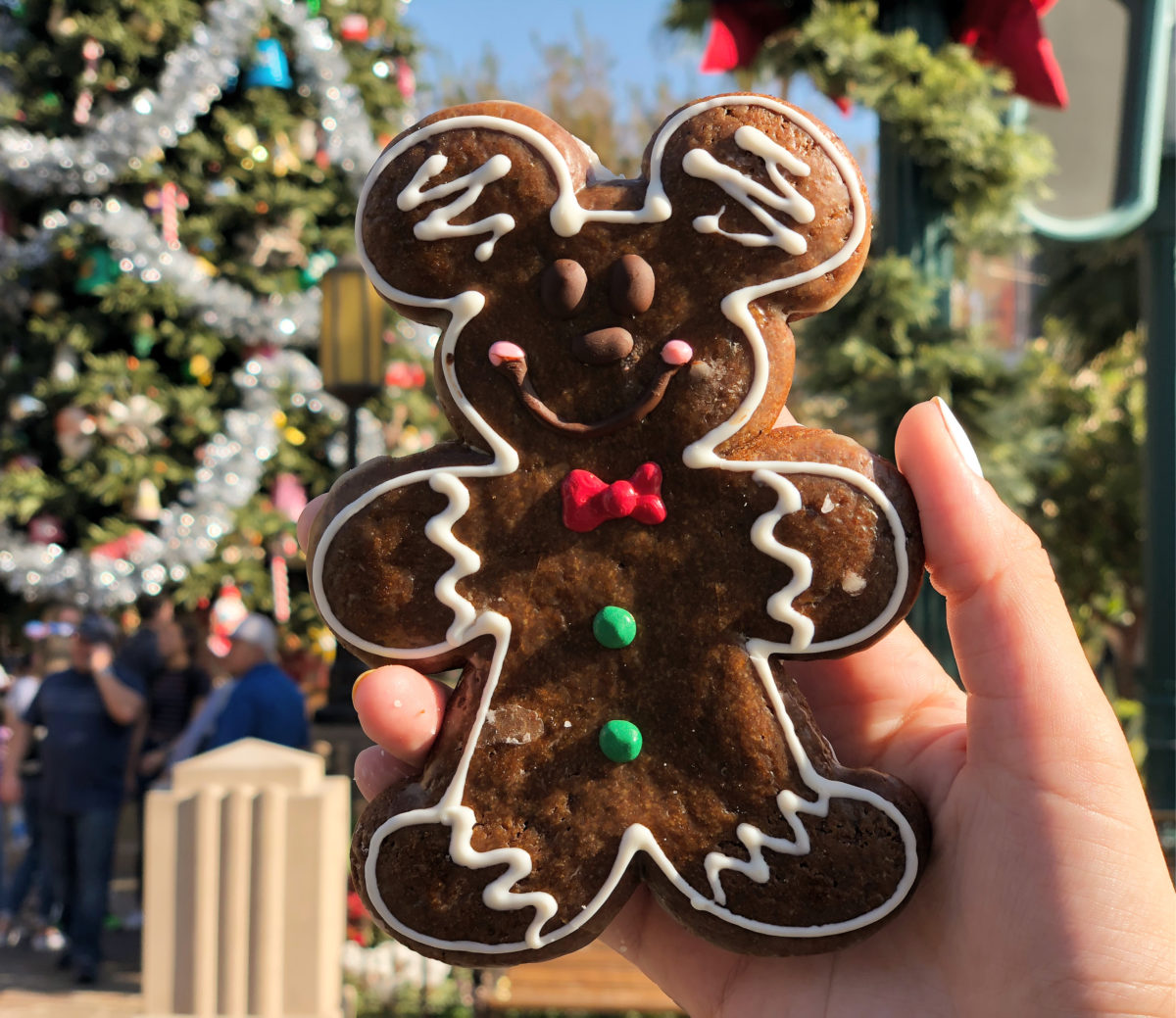 A hand holds a Mickey Mouse-shaped gingerbread cookie from Market House Cookies, adorned with icing details, against the backdrop of a decorated Christmas tree and a blurred scene of people and foliage.