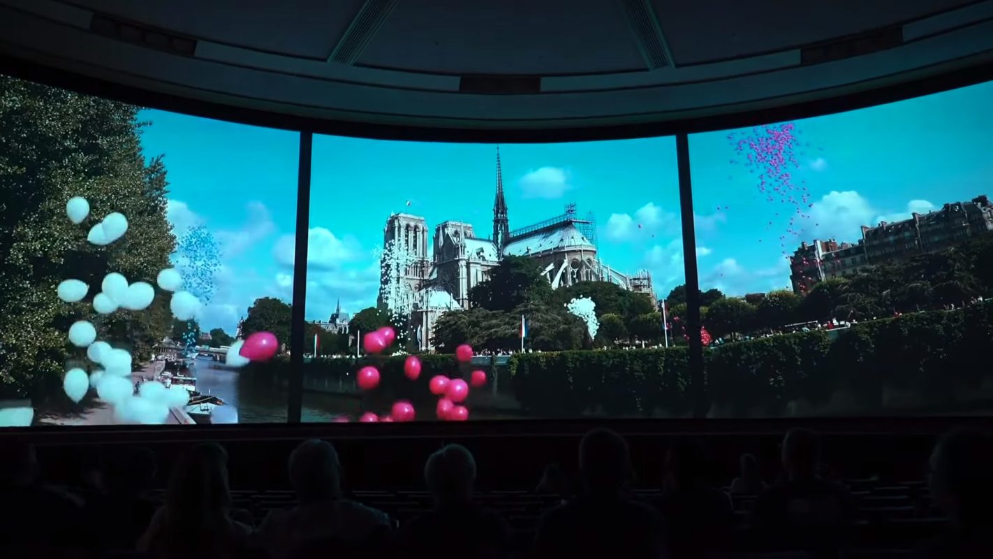 Large screen displays an image of Notre Dame Cathedral with colorful balloons floating in the foreground. People are seated and watching the "Impressions de France" 4K remastered edition.
