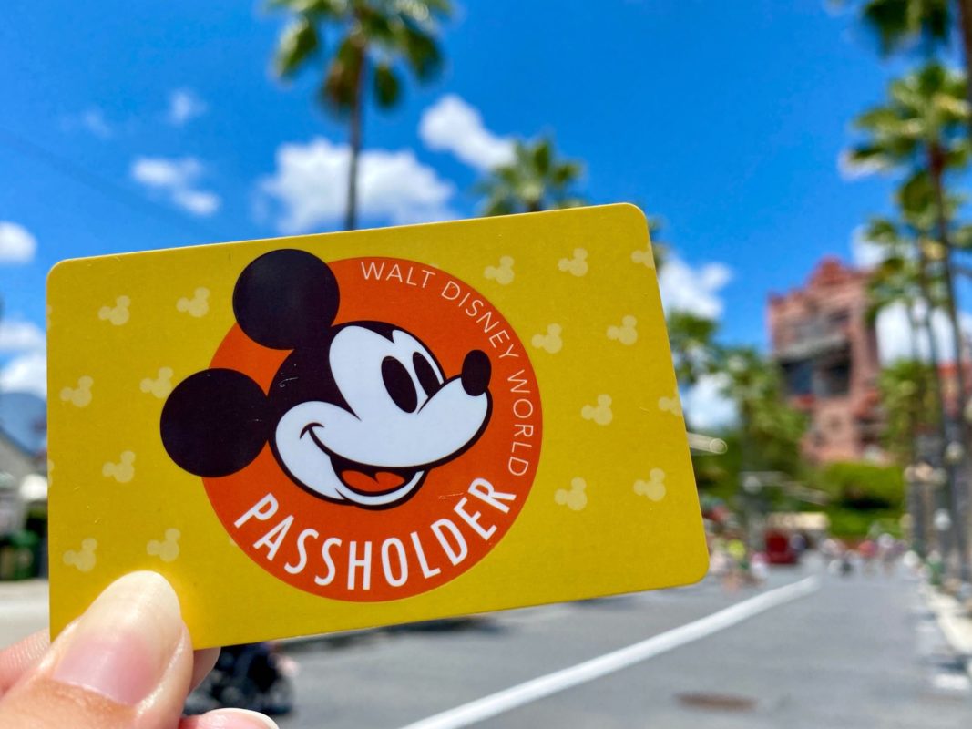 A hand holds a yellow Walt Disney World annual passholder card featuring Mickey Mouse. Palm trees and an amusement park building stand proudly in the background under a clear blue sky.