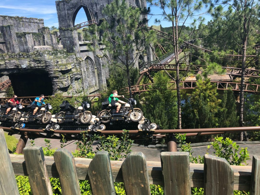People ride a motorcycle-themed coaster through a forested park, enjoying the universal virtual queue on a sunny day.
