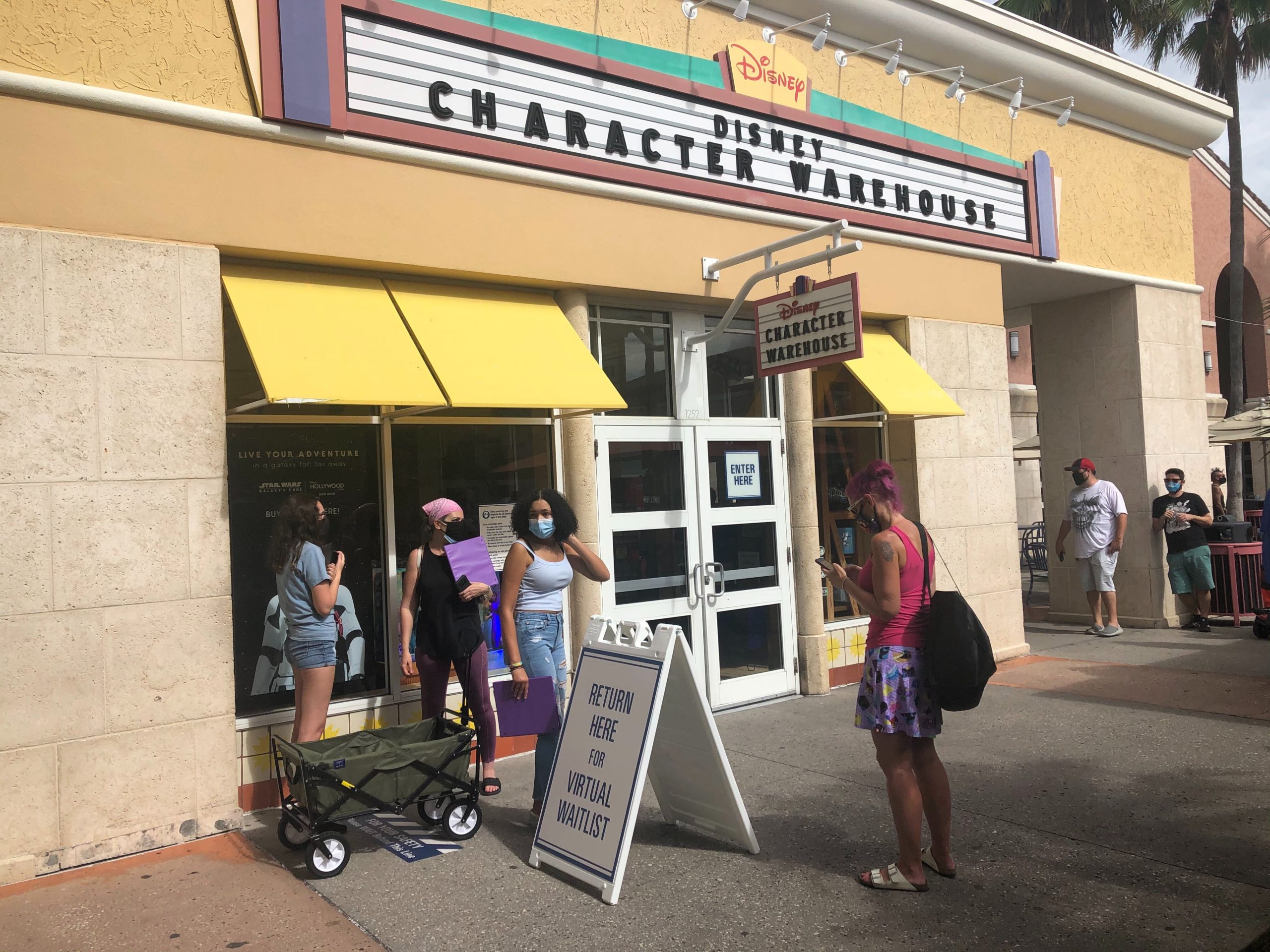 People stand outside Disney Character Warehouse, some wearing masks. A sign directs visitors to "Return Here for Virtual Waitlist." A person with a cart and a stroller are also visible at this bustling outlet store.