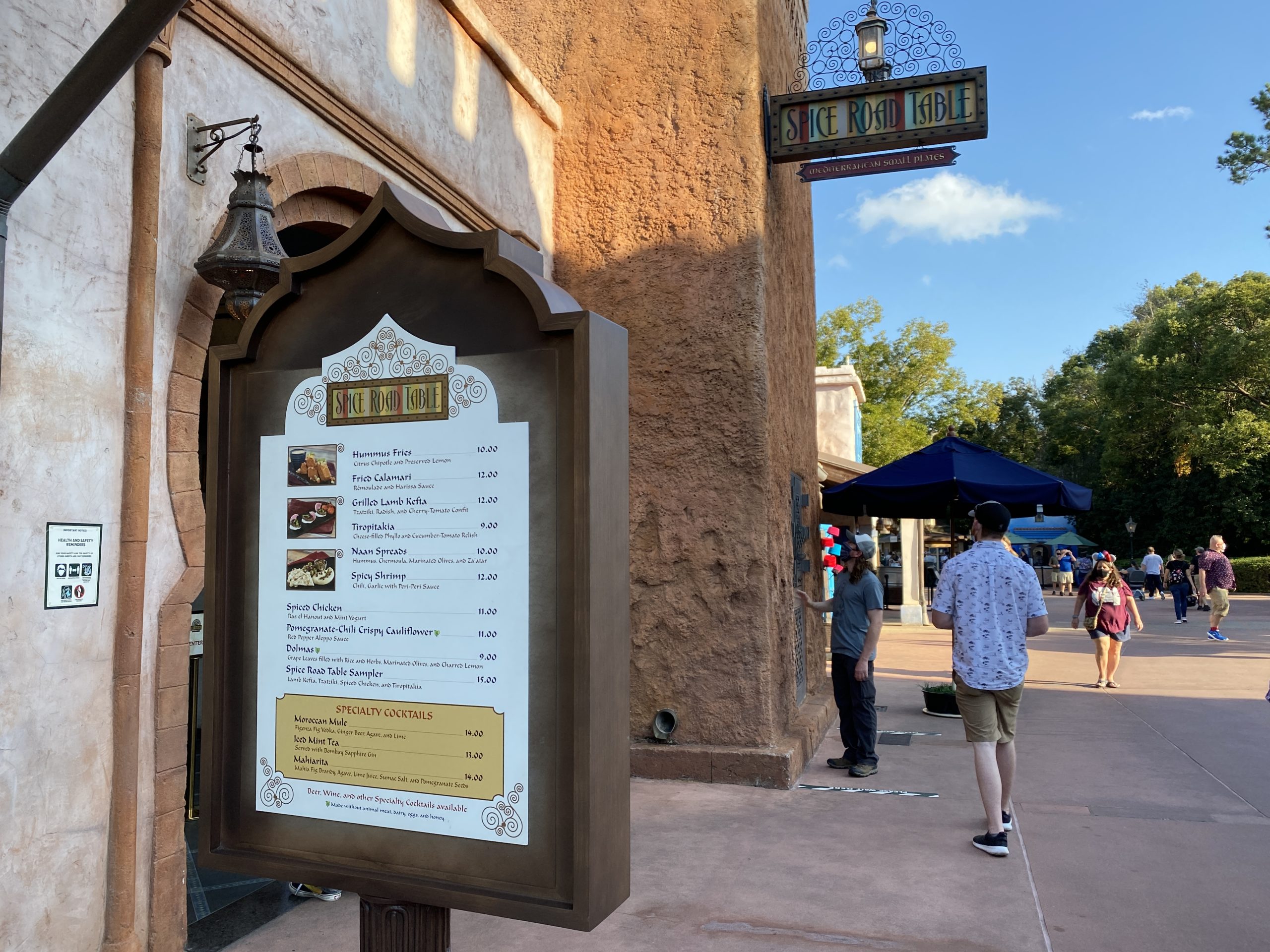 The outdoor restaurant menu display stands gracefully against a textured wall, basking in the sun as a few people stroll by. It almost feels like an Epcot photo report moment, capturing the vibrant blend of culinary choices and leisurely ambiance.