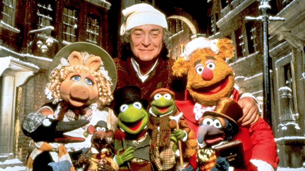 The Muppet characters, dressed in Victorian-style clothing, stand outside a snow-covered building alongside a person in a Scrooge-like outfit, evoking the timeless charm of "The Muppet Christmas Carol."