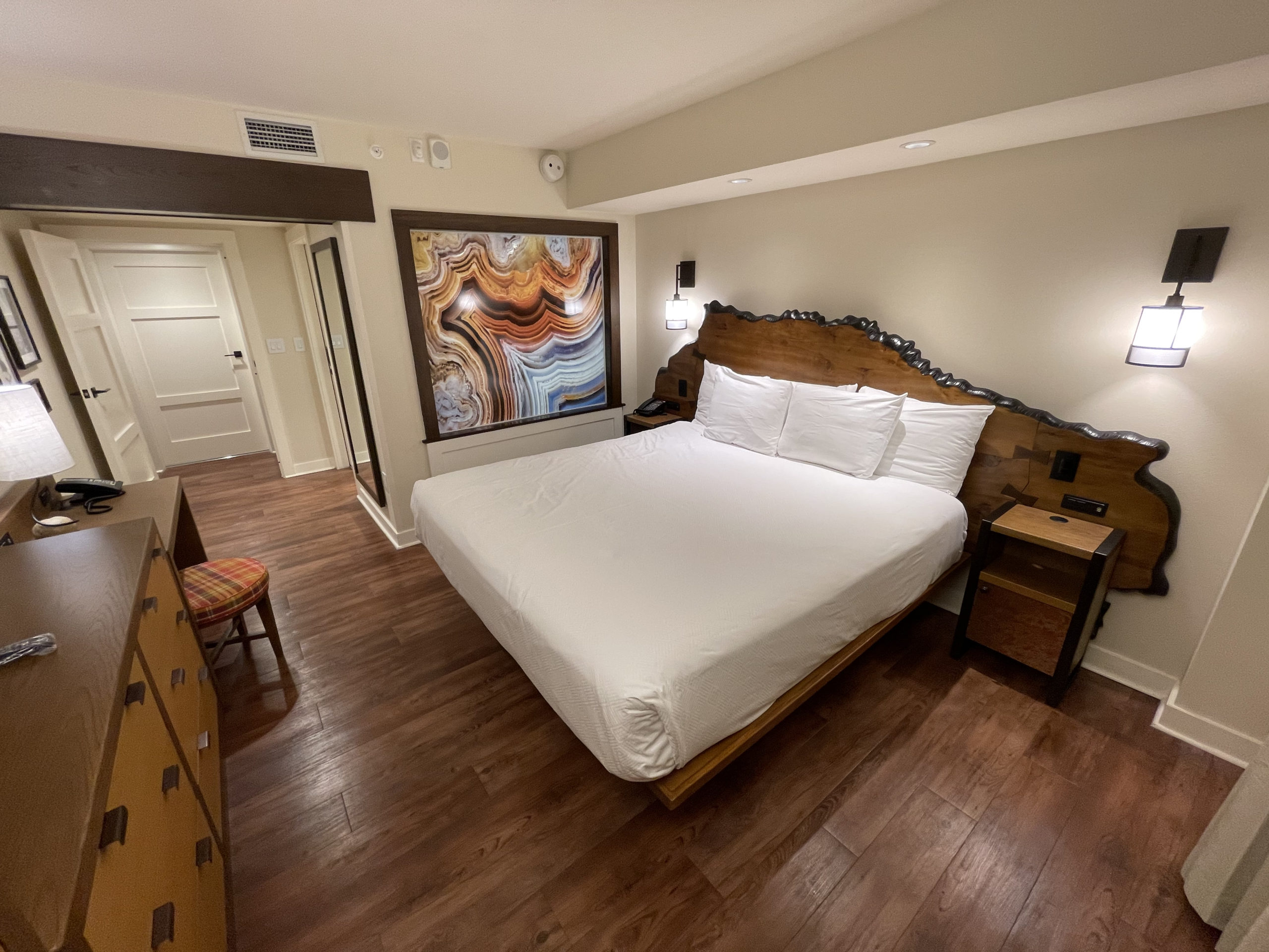 A hotel room with a large bed, wooden furniture, a desk with a lamp, a wall-mounted artwork, and a wooden floor.