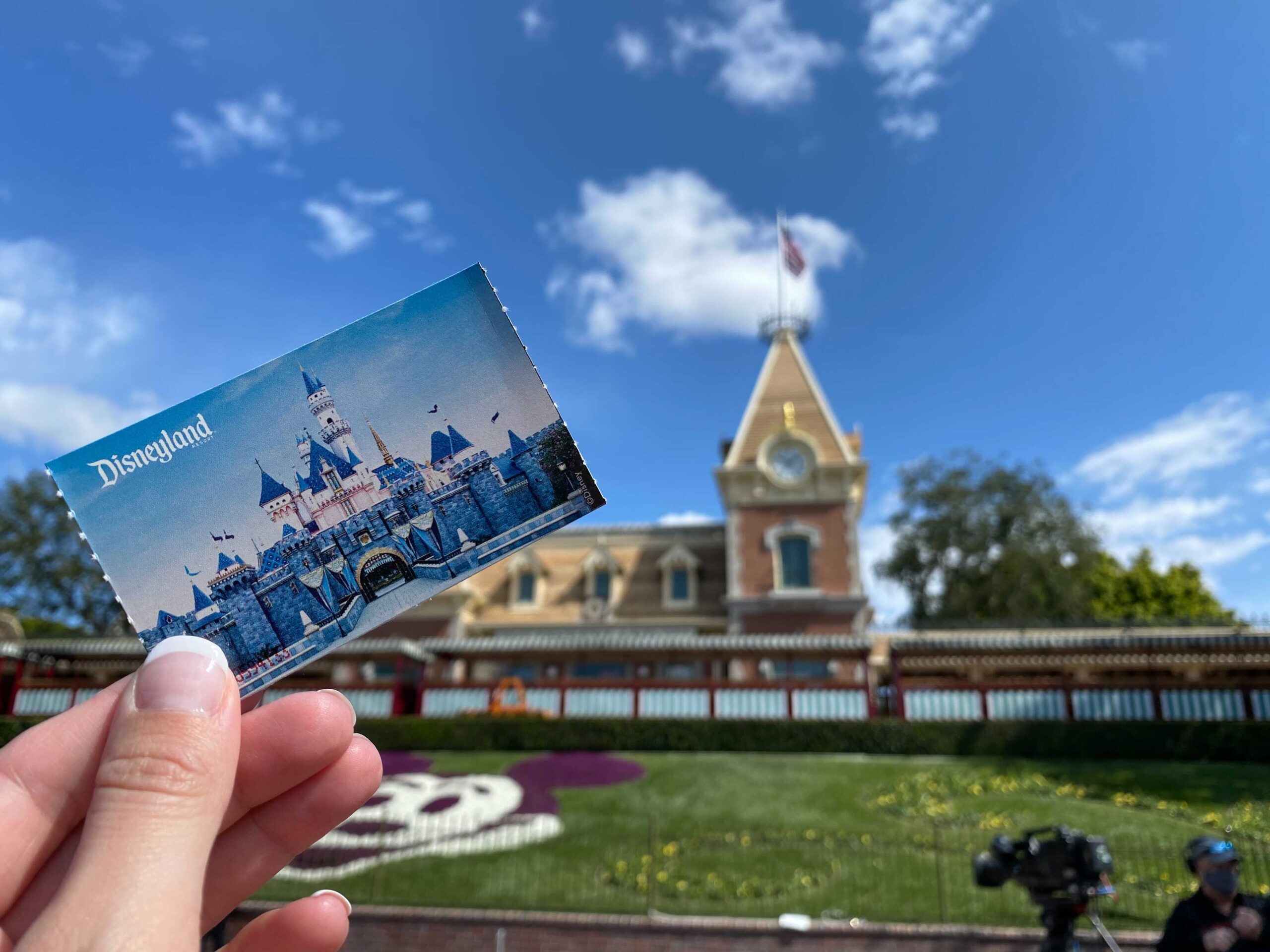 A hand displays a Disneyland residents ticket against the entrance backdrop, showcasing the iconic clock tower and vibrant floral Mickey Mouse design.