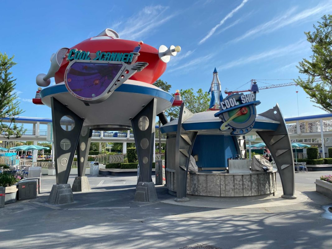 Futuristic drink stand Cool Ship and Cool Scanner mist spray at Magic Kingdom with space-themed designs and a red spaceship on top in a sunny outdoor area, trees, and umbrellas.