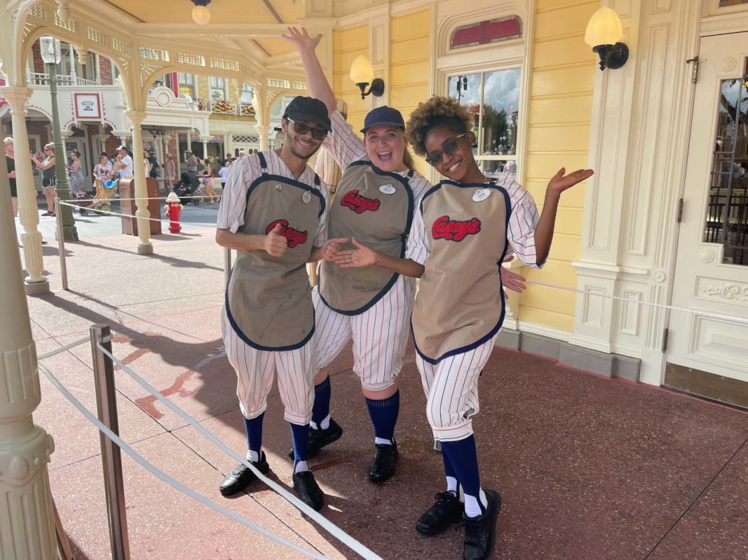 Three people dressed in vintage baseball uniforms pose happily at Casey's Corner, their smiles brightening in front of a yellow building at an amusement park.
