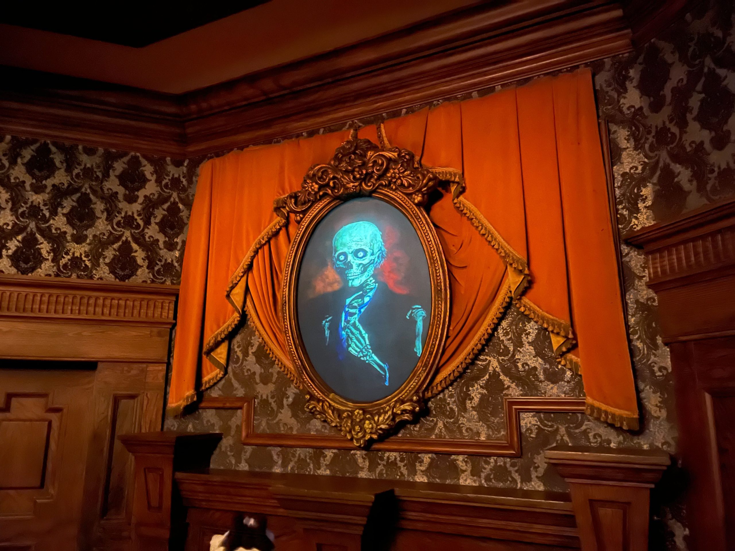 An oval portrait of a skeletal figure, known as the Master Gracey Portrait, is displayed in an ornate gilded frame, set against a backdrop of orange drapery and dark patterned wallpaper.