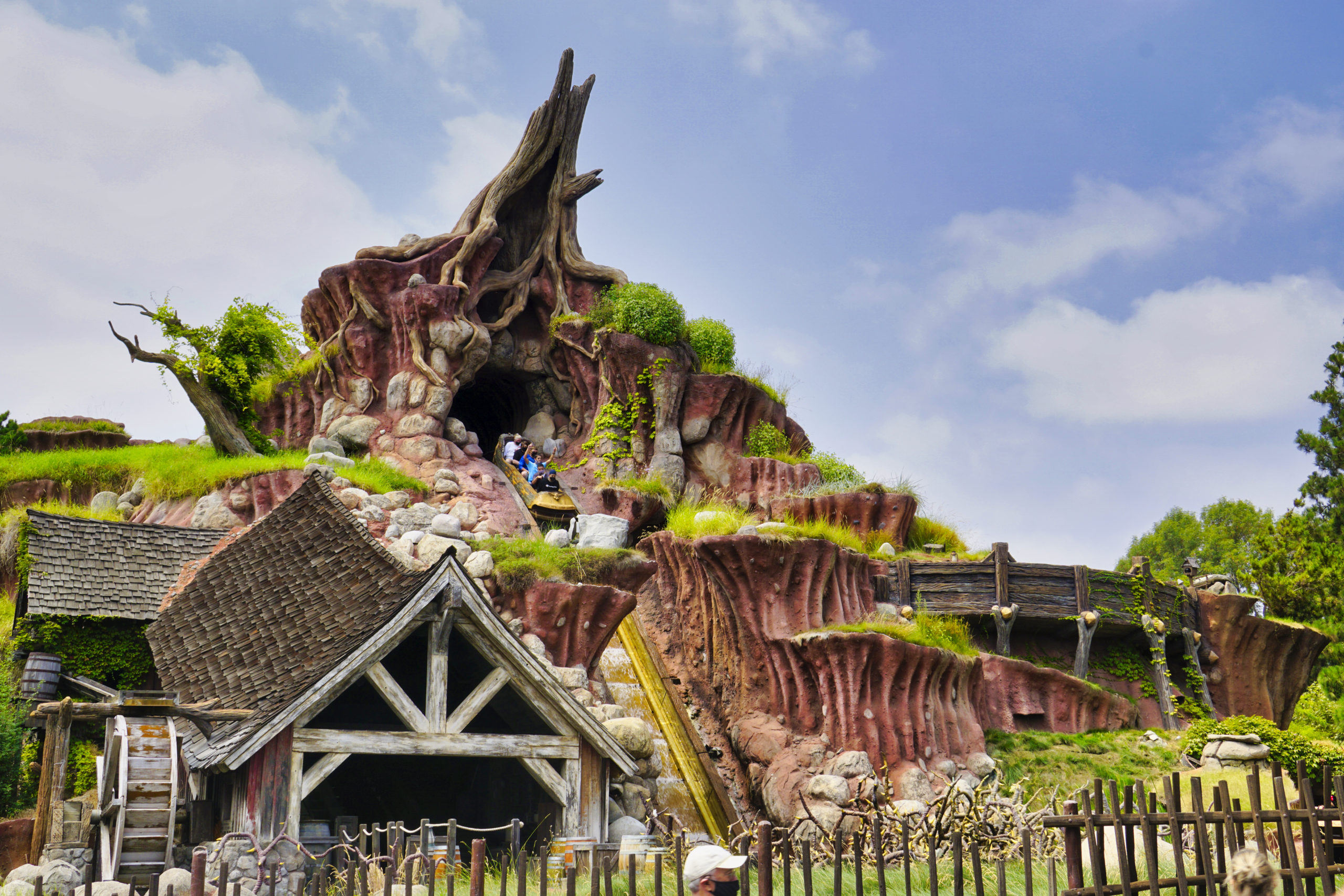 Disneyland's Splash Mountain, featuring a rocky mountain facade, a millhouse, wooden fencing and lush greenery under a blue sky.
