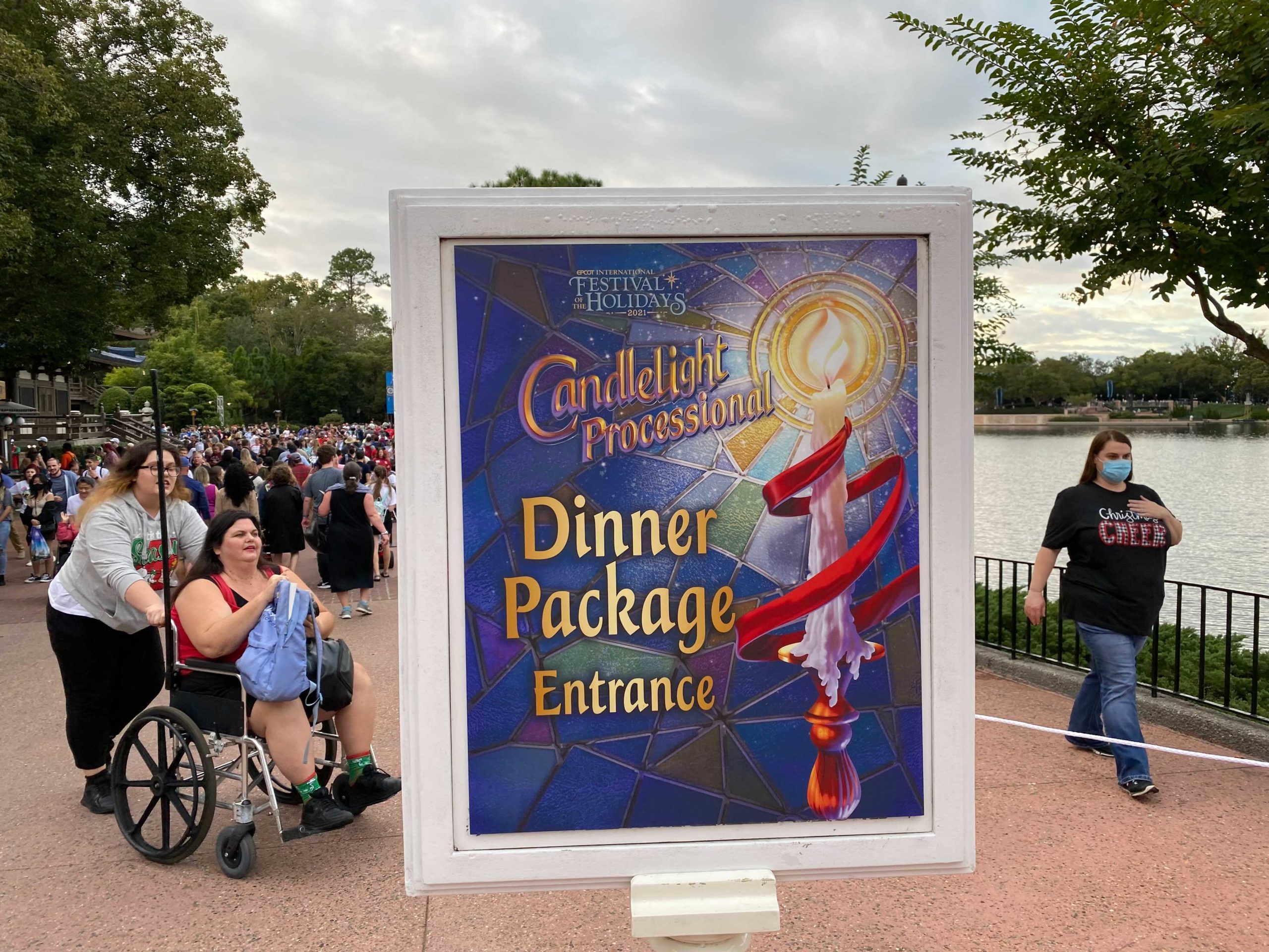Sign for "Candlelight Processional Dinner Package Entrance" at Epcot, with people in the background. A woman in a wheelchair and a masked woman stand nearby on a cloudy day.