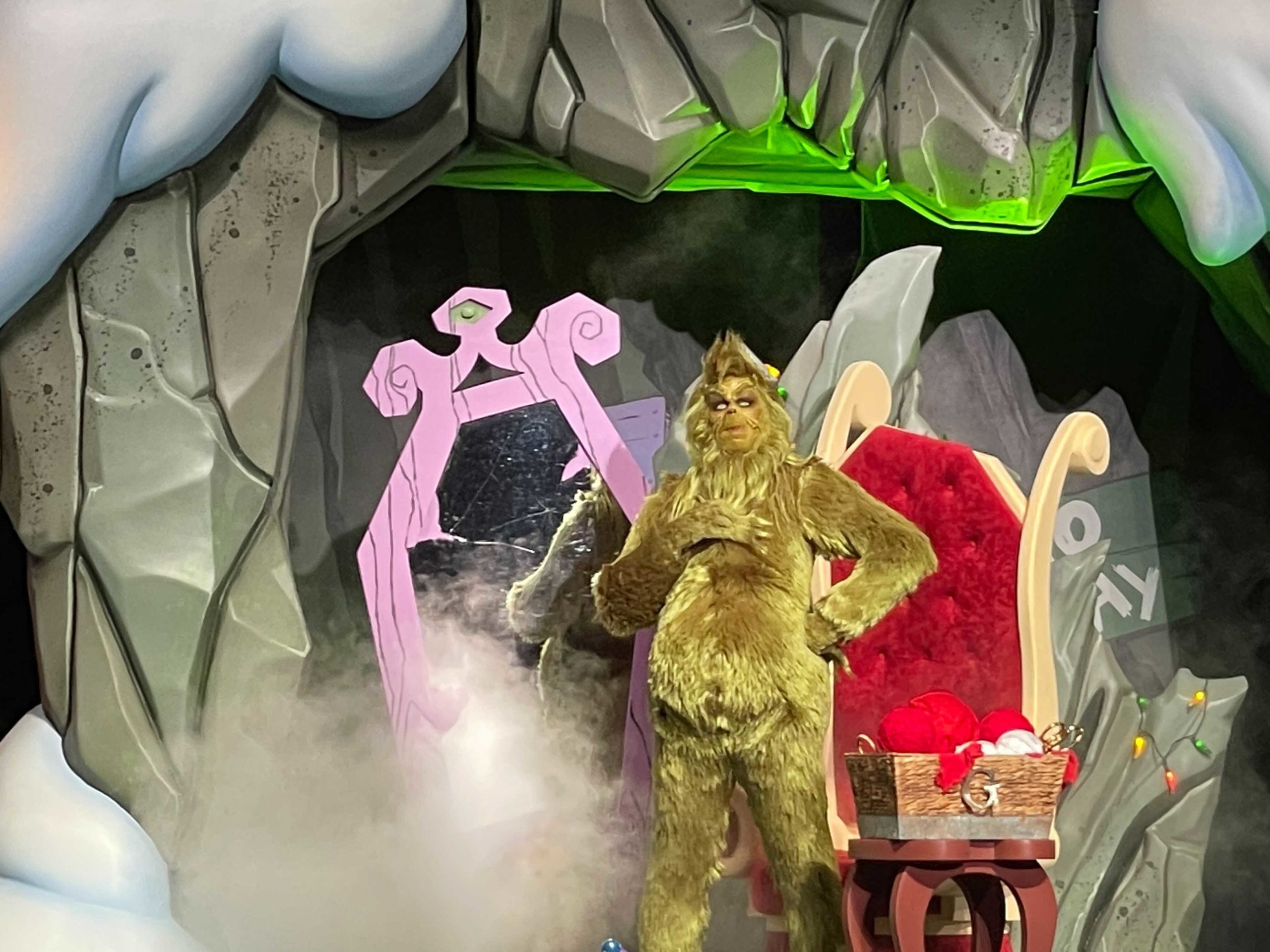 A person in a Grinch costume stands confidently in front of a festive backdrop that includes a large pink mirror and a plush red chair, with mist rising around them. The maskless Grinch seems to embrace the spirit of Grinchmas, adding an extra layer of whimsy to the scene.