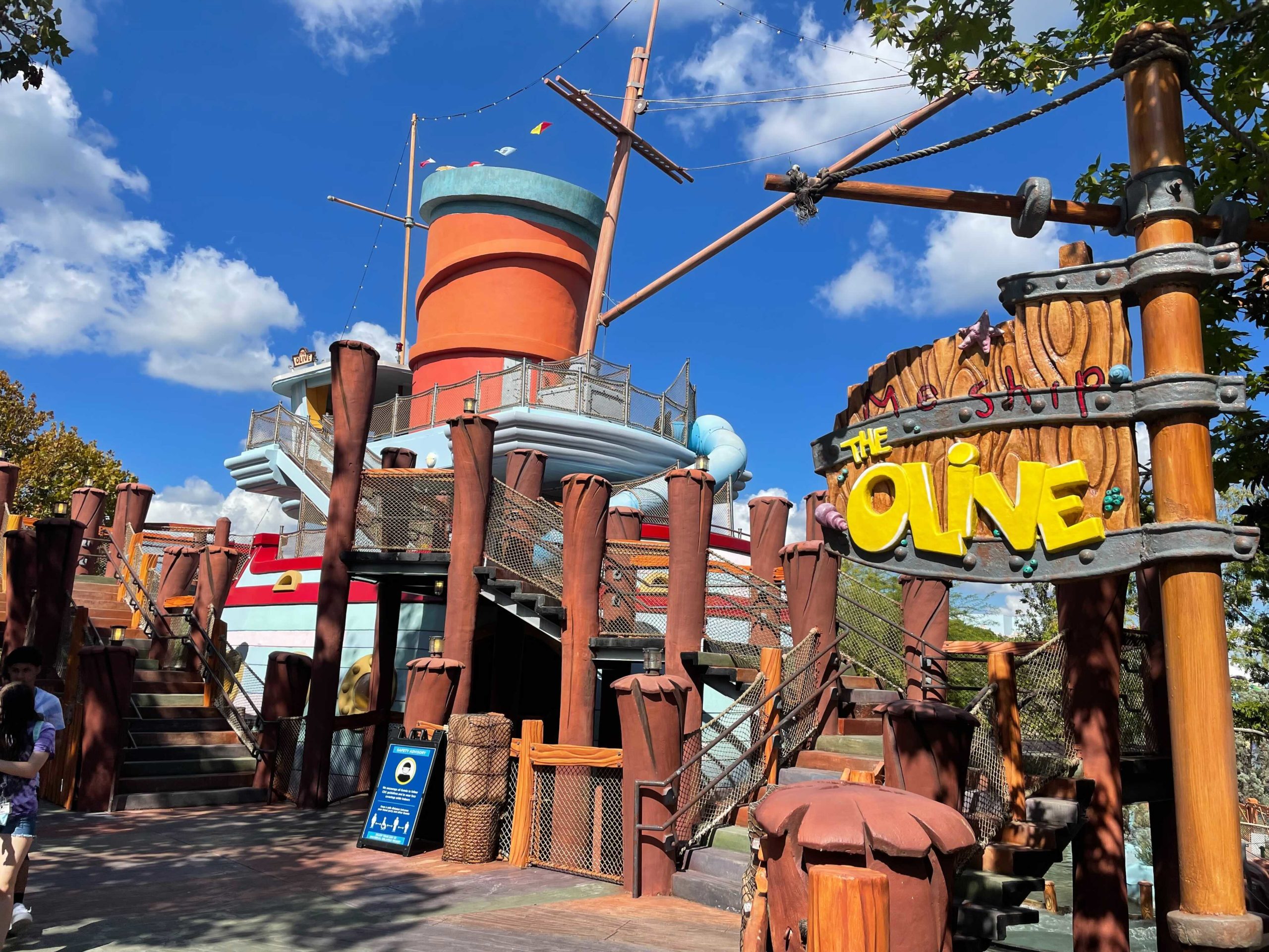 An outdoor play area featuring a large, colorful structure resembling a ship with "Me Ship The Olive" sign. The ship has a red stack, wooden posts, and netted areas. Trees and blue sky are in the background.