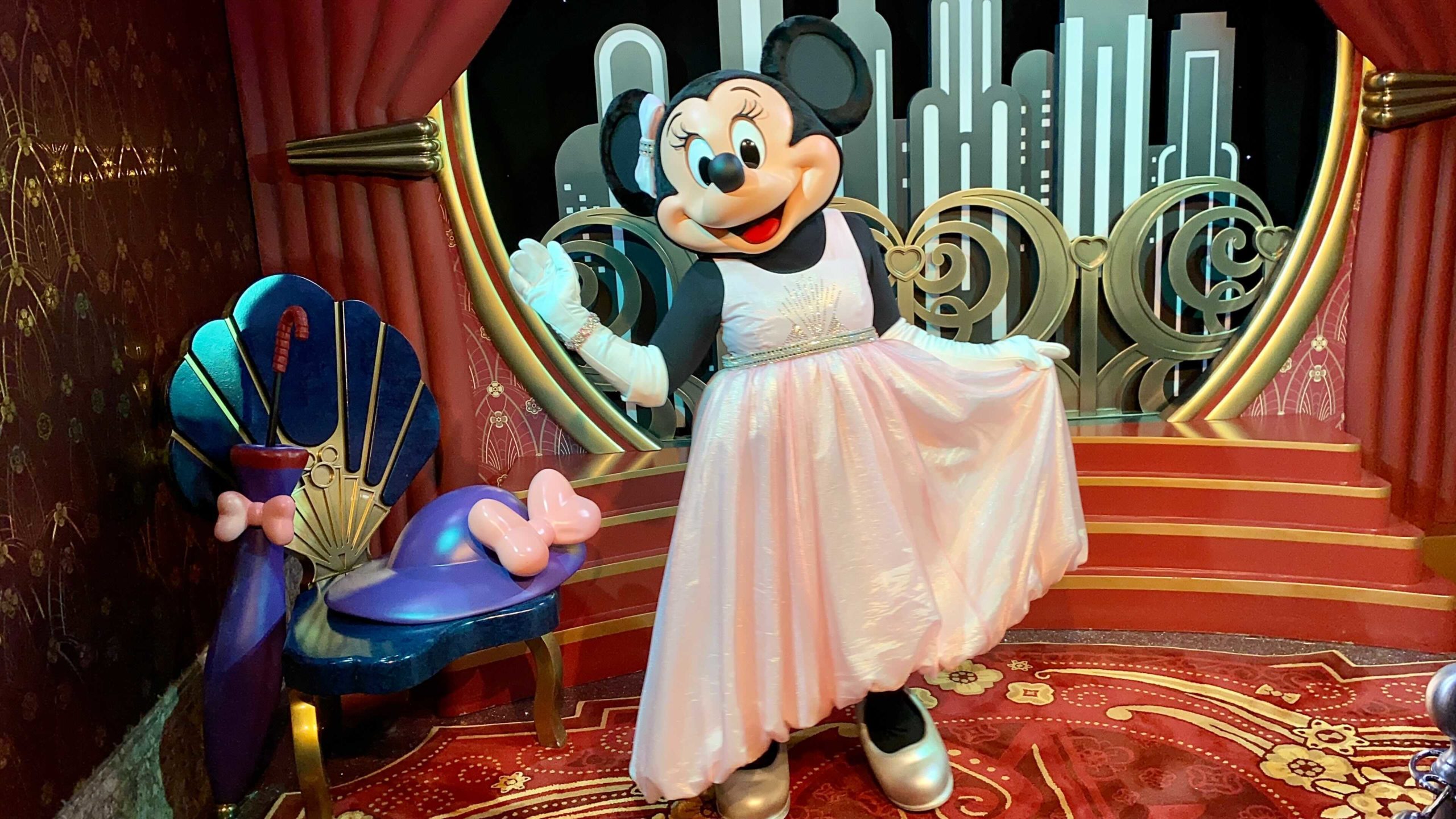 Minnie Mouse, elegantly dressed in white, poses on a red carpeted stage at Disney's Hollywood Studios, with a stunning cityscape backdrop.