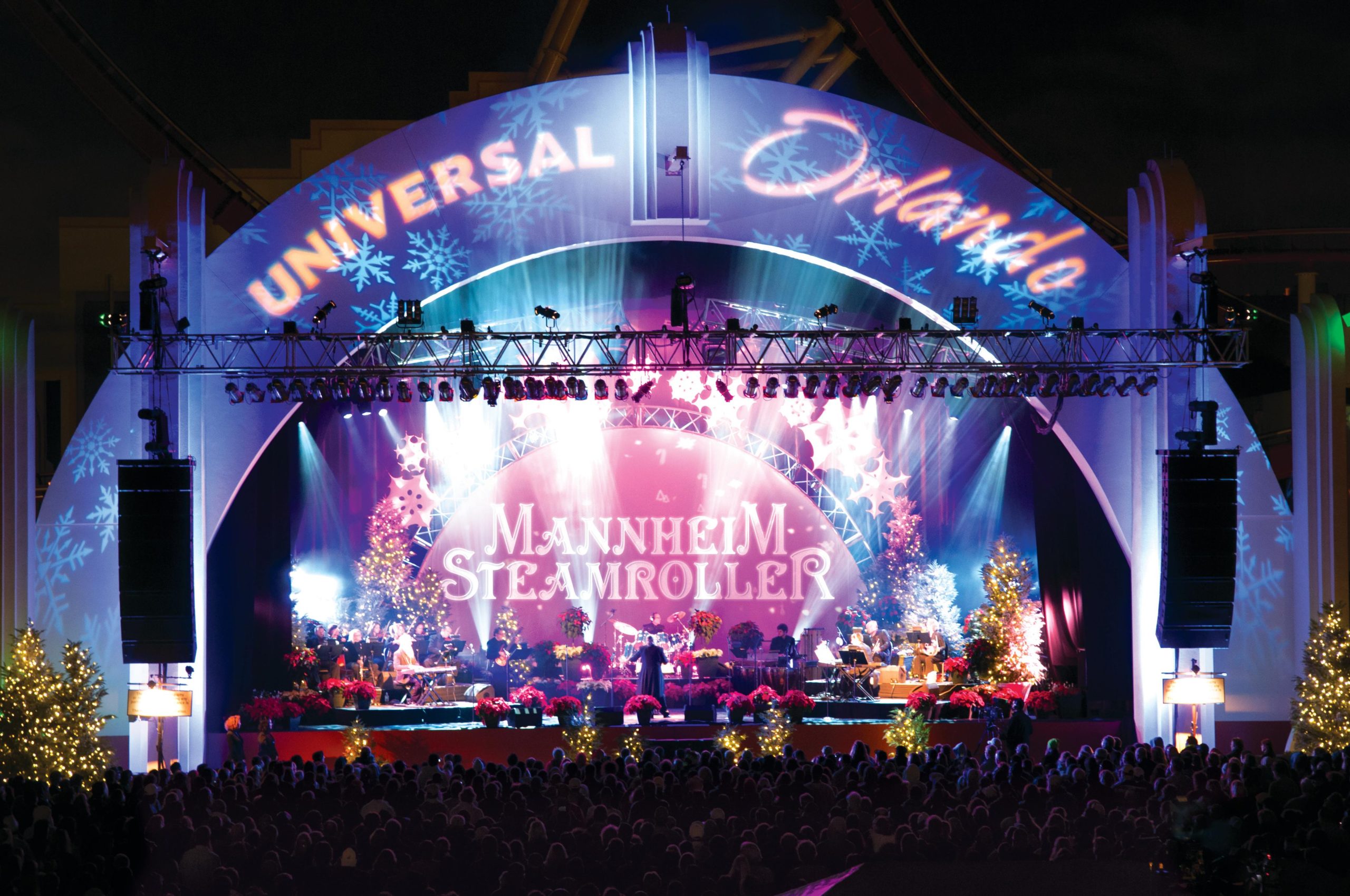 A nighttime concert at Universal Orlando dazzles with Mannheim Steamroller, featuring colorful stage lights and festive holiday decorations, including Christmas trees and snowflakes.