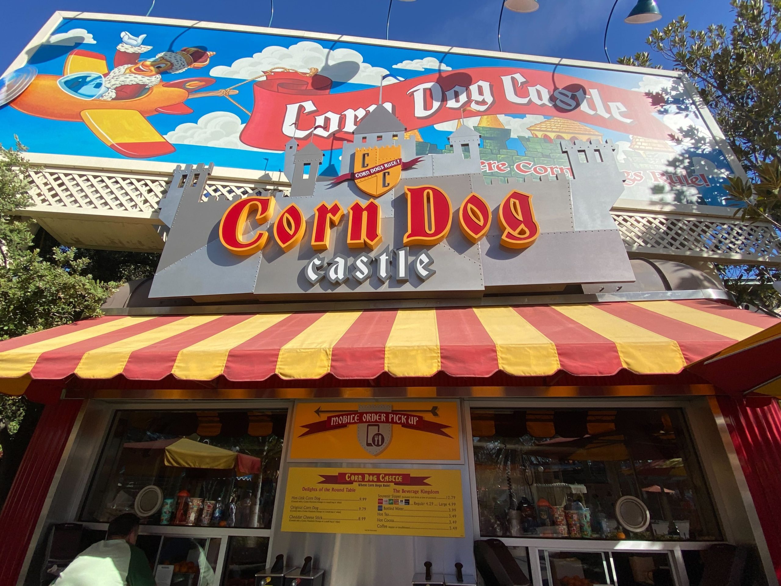 Corn Dog Castle food stand, with its striped awning and vibrant signage, now features a green corn dog as the star attraction on the colorful billboard above.