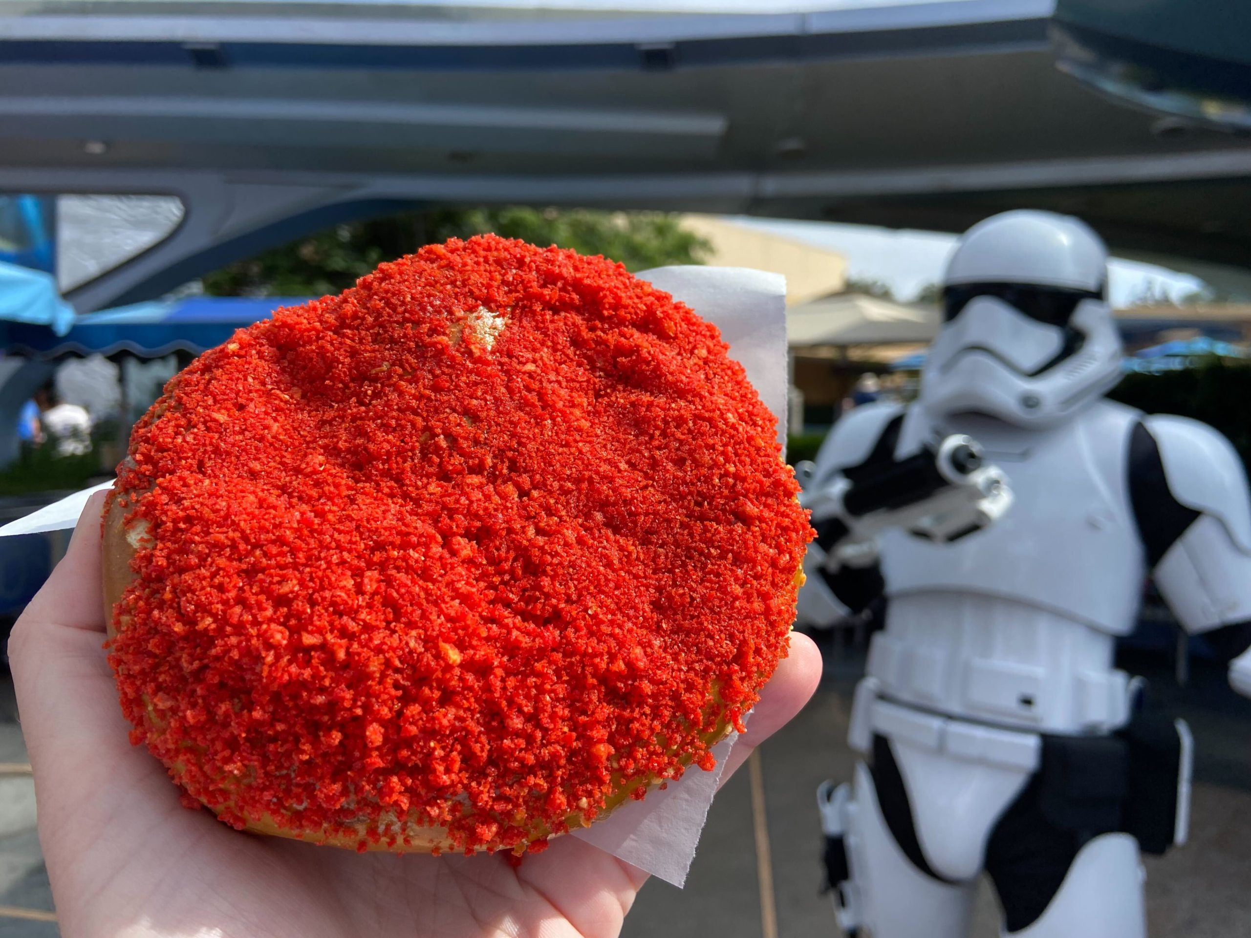 A hand holds a pastry covered in red crumbs, resembling a Supernova Pretzel, with a stormtrooper in the background at an amusement park.
