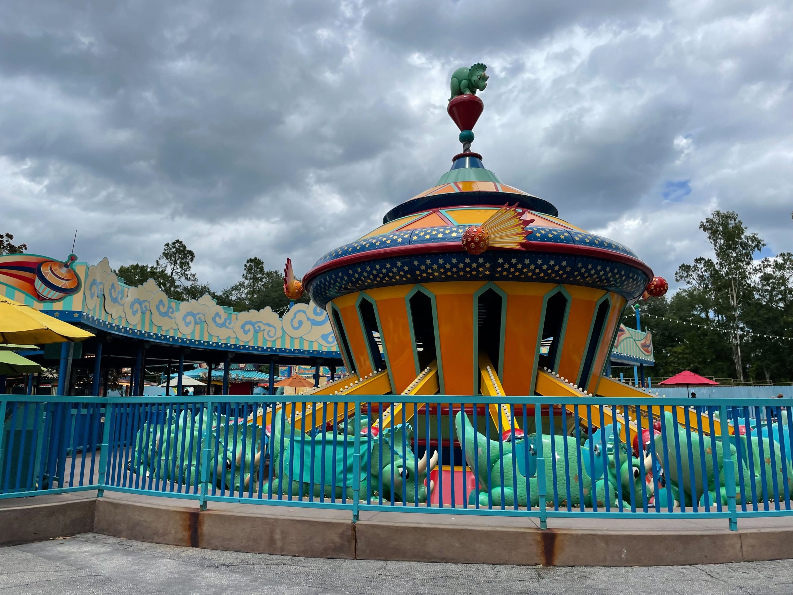 Colorful amusement ride featuring triceratop seats, encircled by a blue fence under a cloudy sky, freshly spruced up during its recent refurbishment.