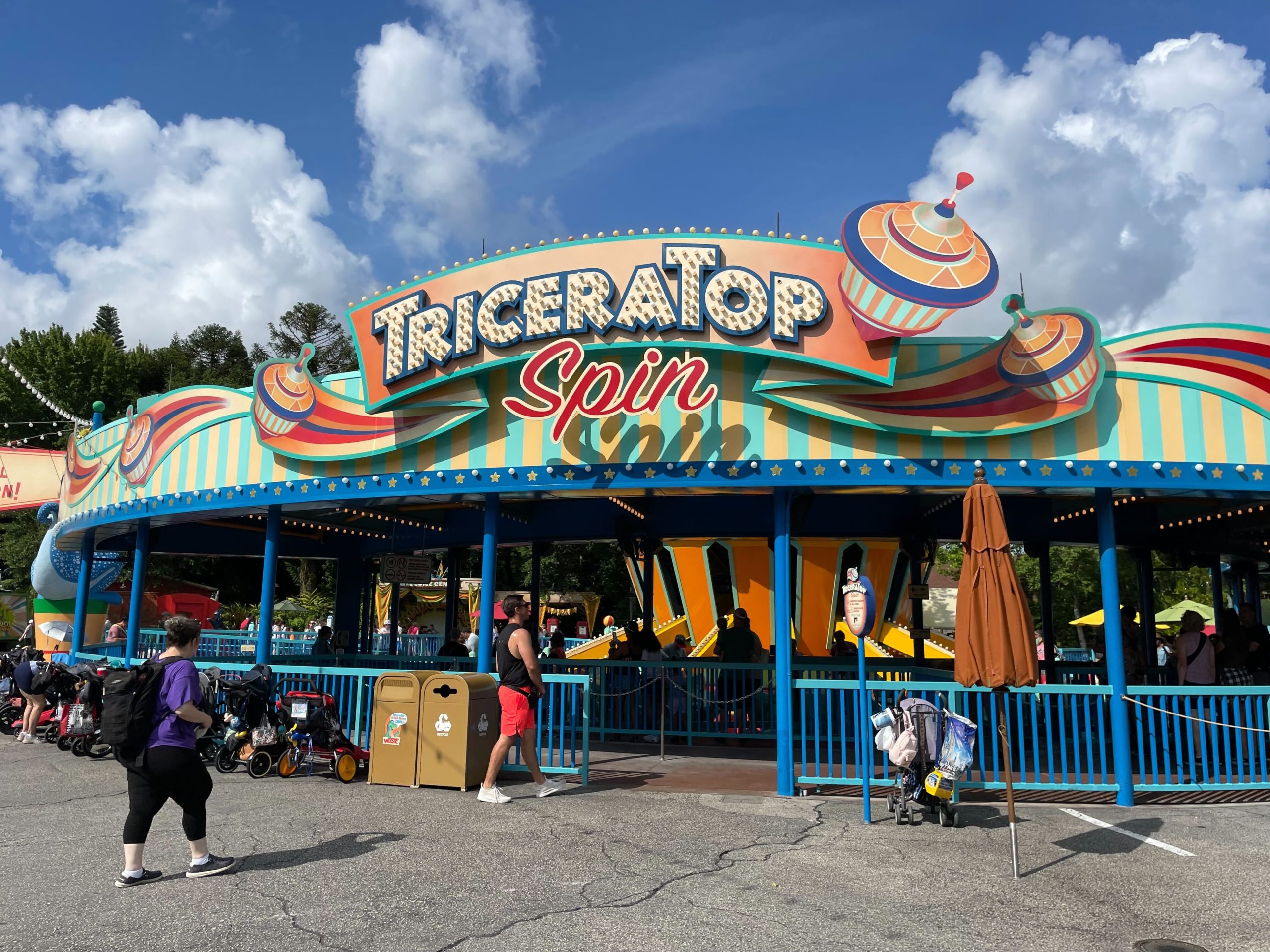 The vibrant "TriceraTop Spin" ride, freshly enhanced by a recent refurbishment, shines with spinning tops on the sign. A lively scene unfolds as people and strollers gather outside on a sunlit day.
