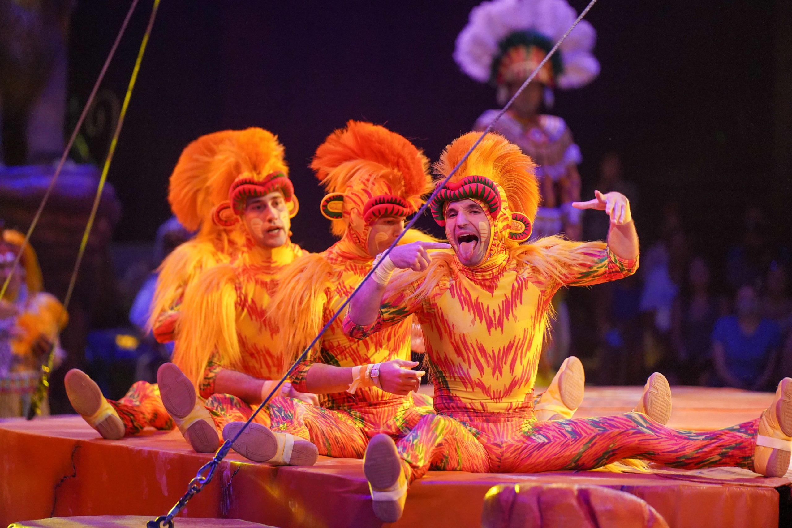 Four performers in vibrant orange and yellow costumes sit on a stage. One performer sticks out their tongue and gestures animatedly. The background is dimly lit with a standing figure barely visible. It's clear that the Festival of the Lion King has finally returned, dazzling audiences once again.