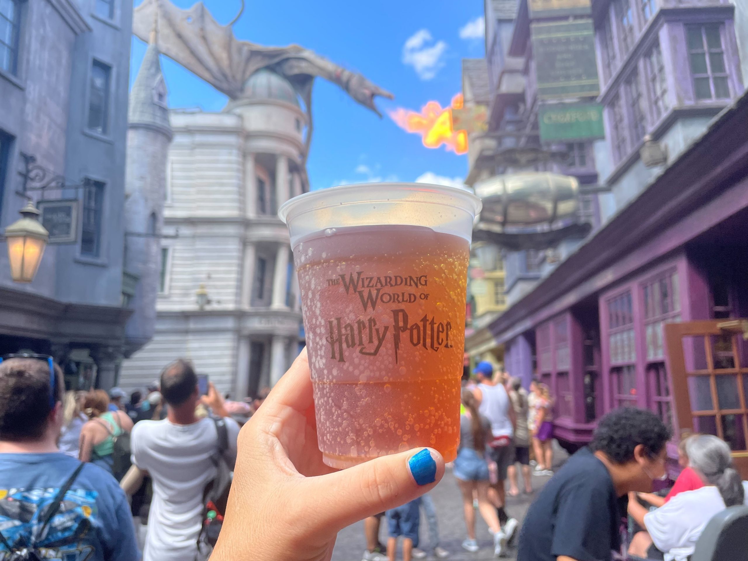 A person holds up a frosty Butterbeer in front of Diagon Alley at The Wizarding World of Harry Potter, with a dragon perched on a building in the background, capturing the magic of Universal Orlando Resort.