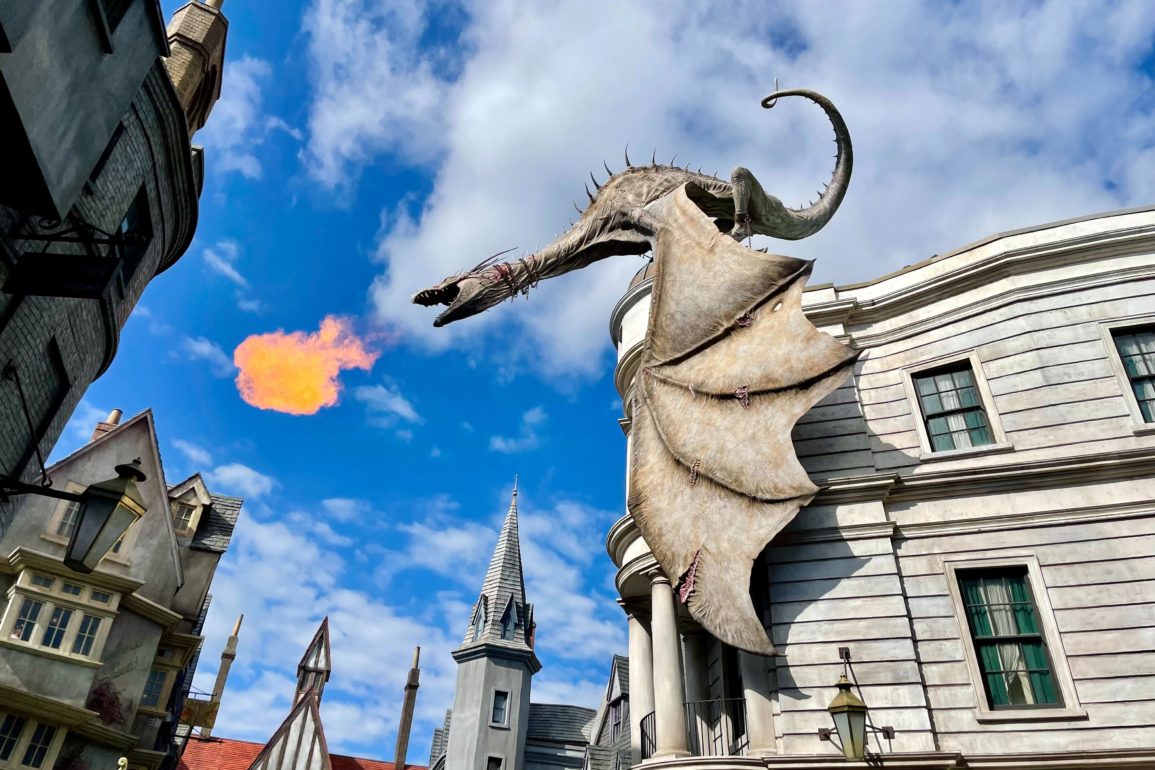 A dragon sculpture on a building facade breathes fire against a backdrop of blue sky and other fantasy-themed buildings, reminiscent of the magical world you can explore with a Universal Orlando Resort ticket.