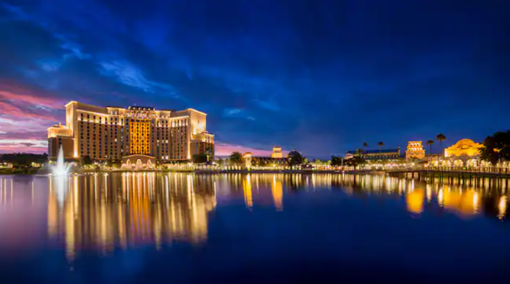 Hotel buildings with illuminated windows are reflected in a calm lake at dusk, beneath a deep blue sky with hints of sunset, while nearby, the ranchos leisure pool offers a serene escape.