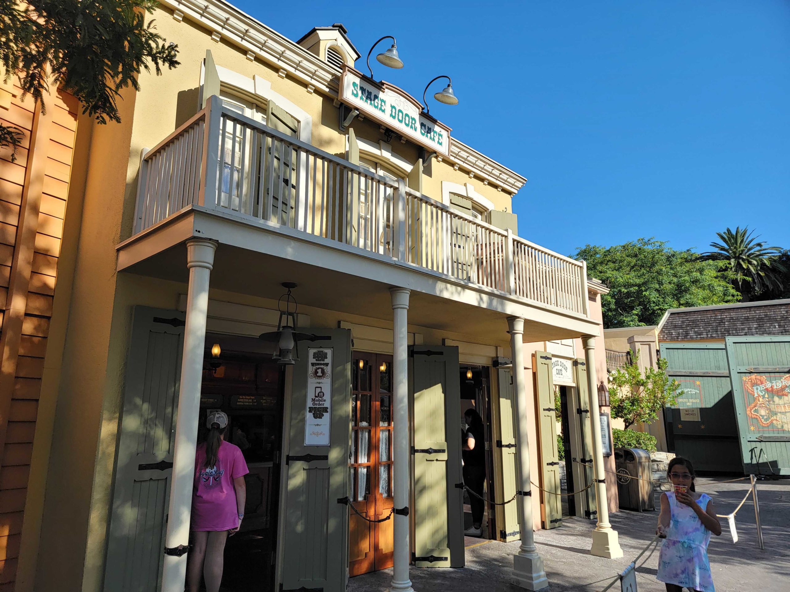 A two-story building with a balcony and a sign saying "Three Door Inn" invites guests with the sweet aroma of strawberry funnel cake wafting through the air. People gather outside, chatting happily as one person enters through a door.