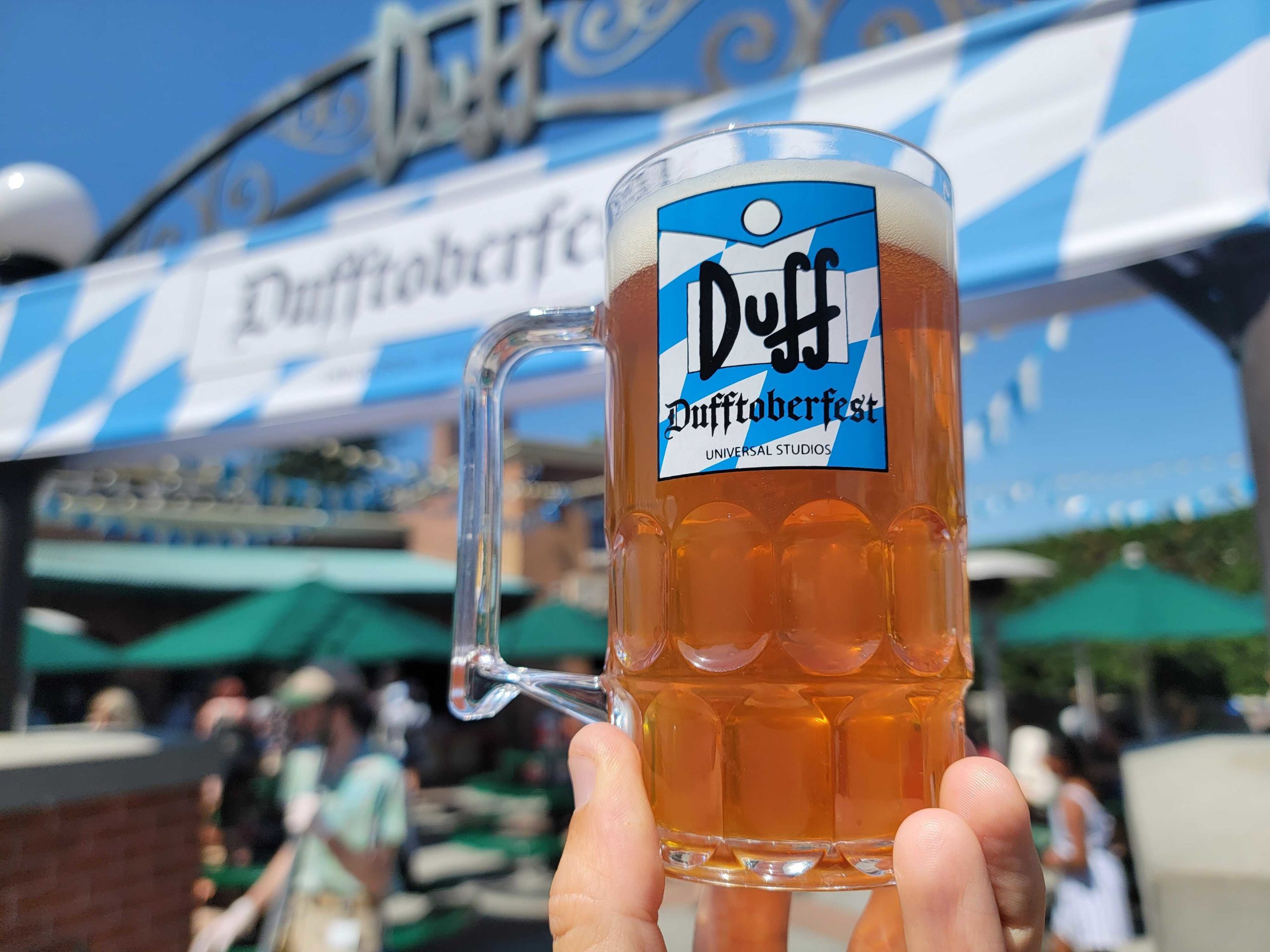 A hand holds a beer mug branded with "Duff"; people and green umbrellas in the background under a sign that proudly announces "Dufftoberfest.