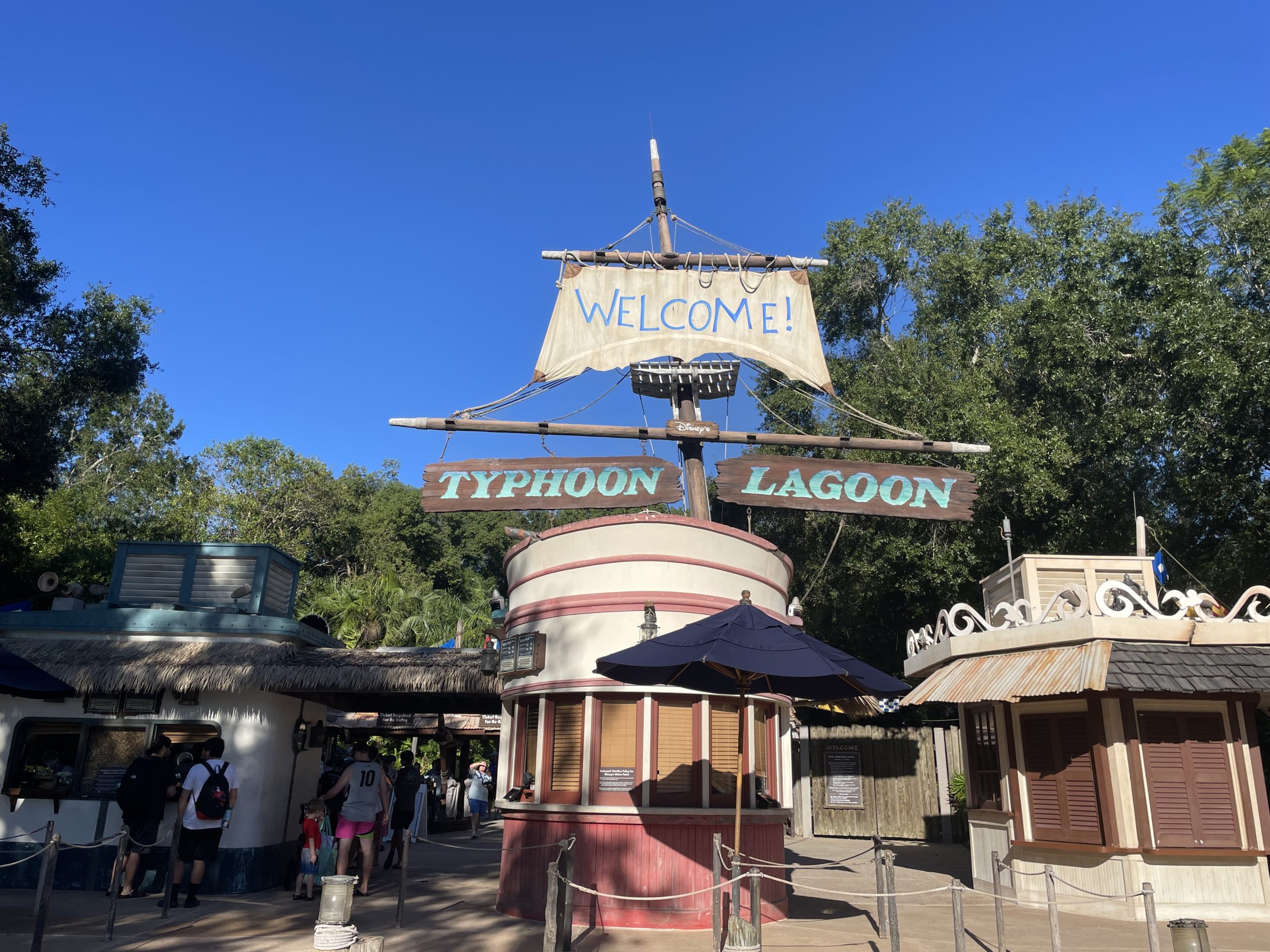 Entrance to Disney’s Typhoon Lagoon with a sign that says "Welcome!" A ticket booth is in the center, surrounded by a few people and lush trees in the background. Annual passholder perks are available for frequent visitors.