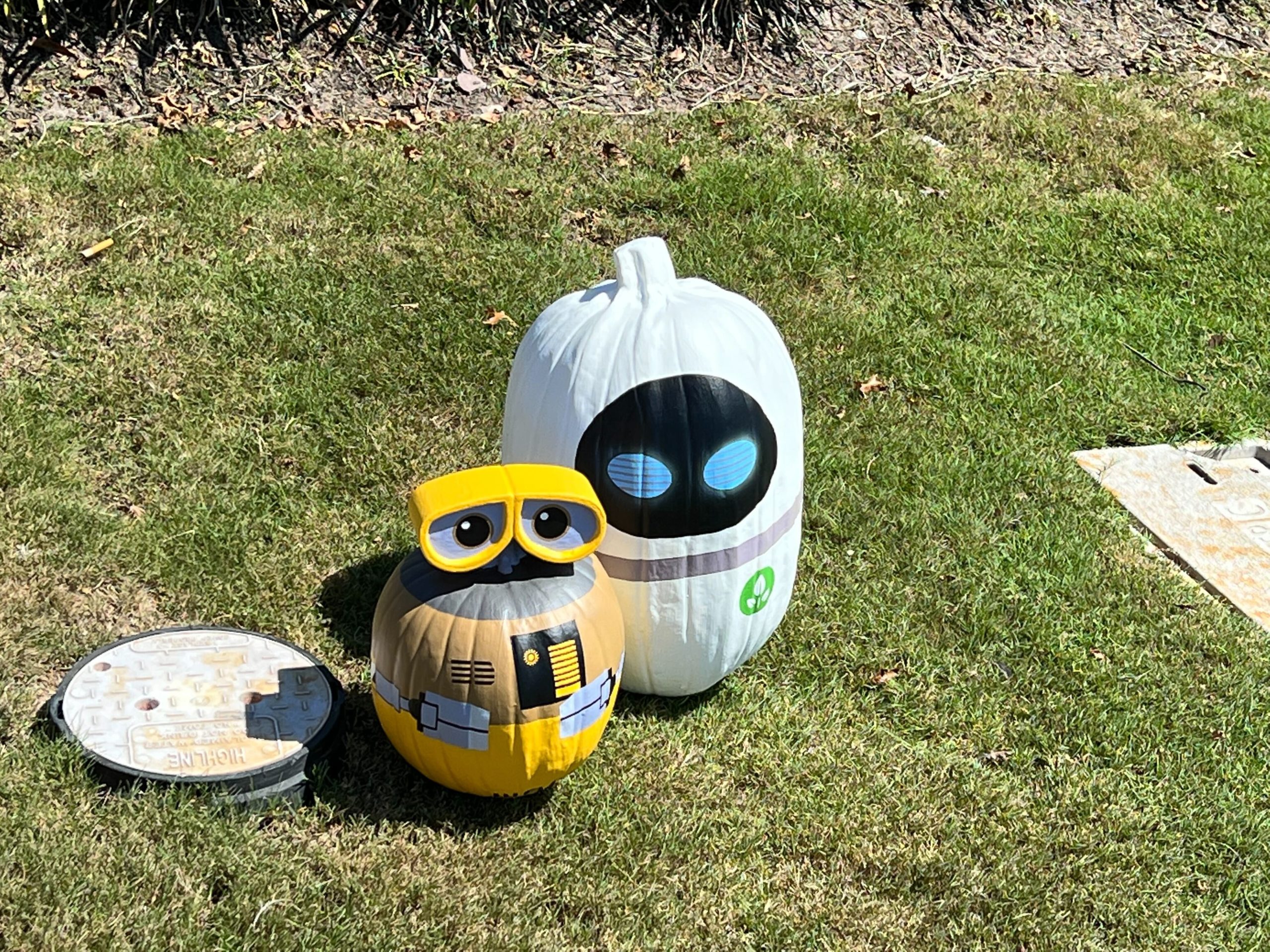 Two pumpkins painted as characters, a yellow and brown one with glasses and a white one with blue eyes, are displayed on grass along with a painted round object, appearing ready for Pluto's Pumpkin Pursuit.