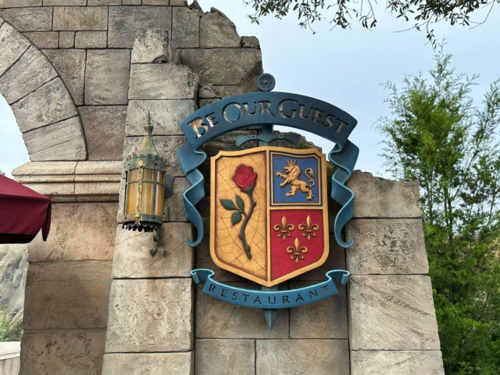 Sign reading "Be Our Guest Restaurant" with a coat of arms featuring a rose and various emblems, mounted on a stone wall, next to an ornate lantern.