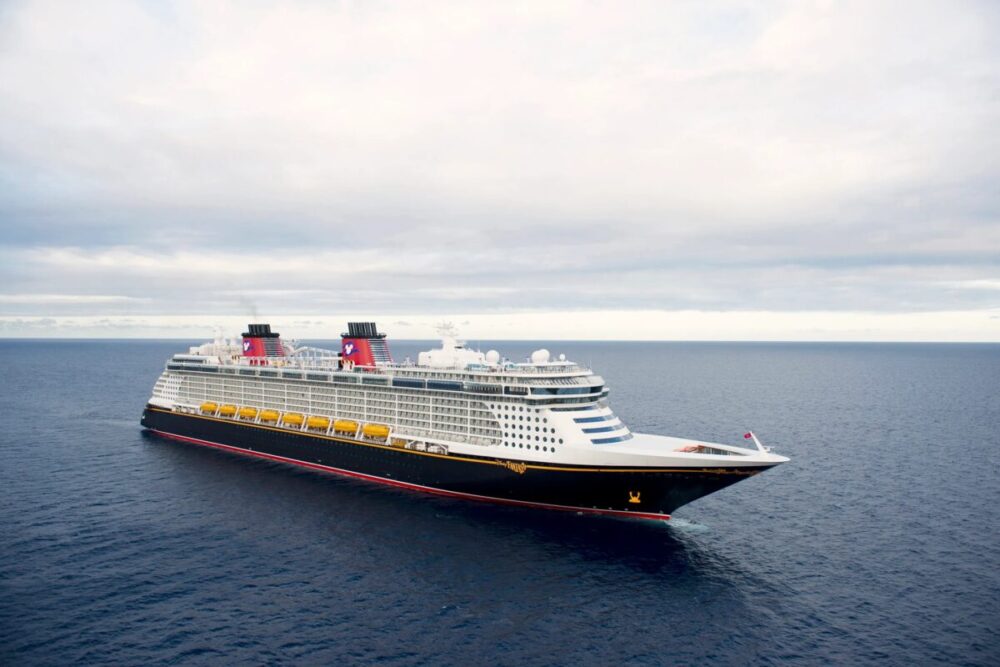 A large Disney Cruise Line ship with distinctive red and black funnels sails on the open sea under a cloudy sky.